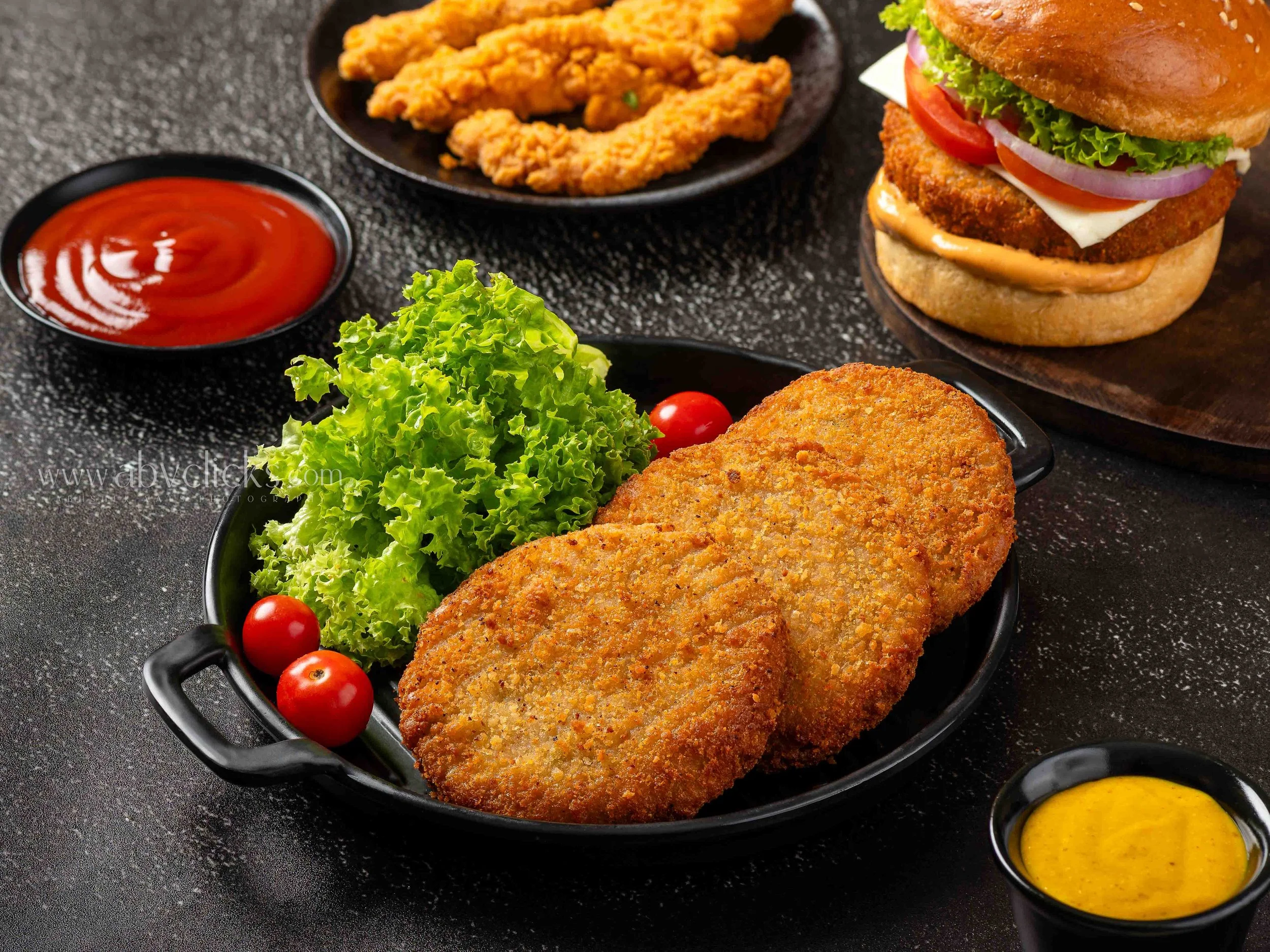 Fried chicken fried steak served with fresh cherry tomatoes, leafy lettuce, and a side of ketchup, along with a large burger with lettuce, tomato, onion, cheese, and a breaded patty in a sandwich bun, and fried chicken tenders with dipping sauce on a