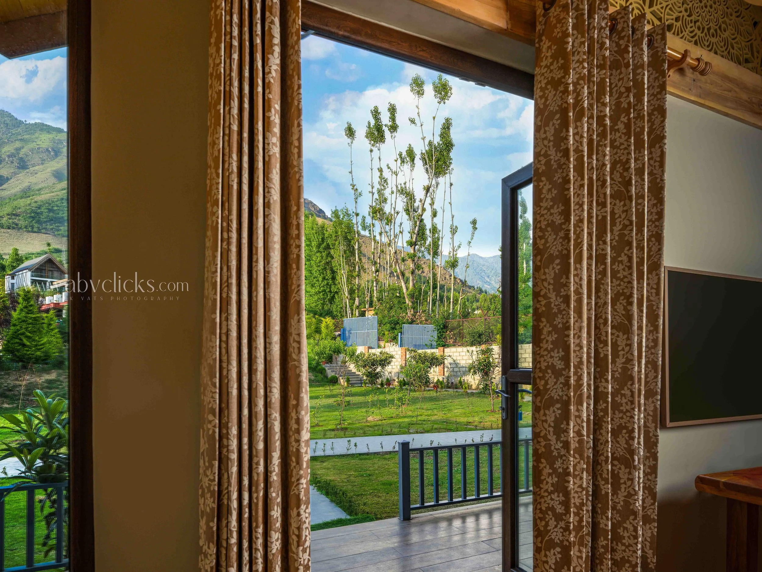 A view through an open sliding glass door shows a grassy yard with small trees, a mountain landscape, and a partly cloudy sky outside. The door is framed by floral patterned curtains.