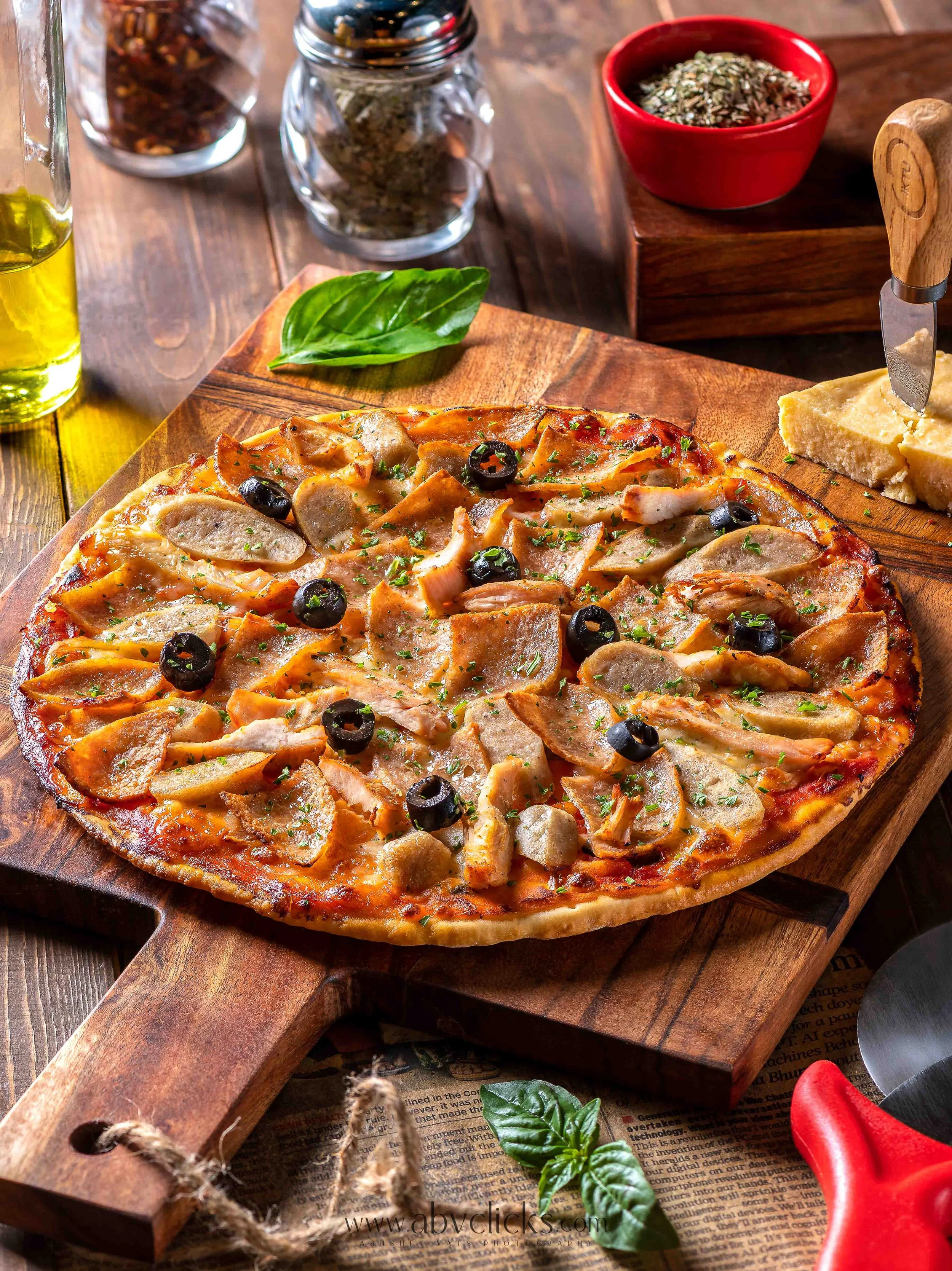 A wooden board with pizza topped with chicken pieces, black olives, herbs, and cheese, placed on a rustic wooden table with basil leaves and a bunch of herbs nearby. Behind the pizza, there are jars of spices, a bottle of olive oil, and a red bowl of