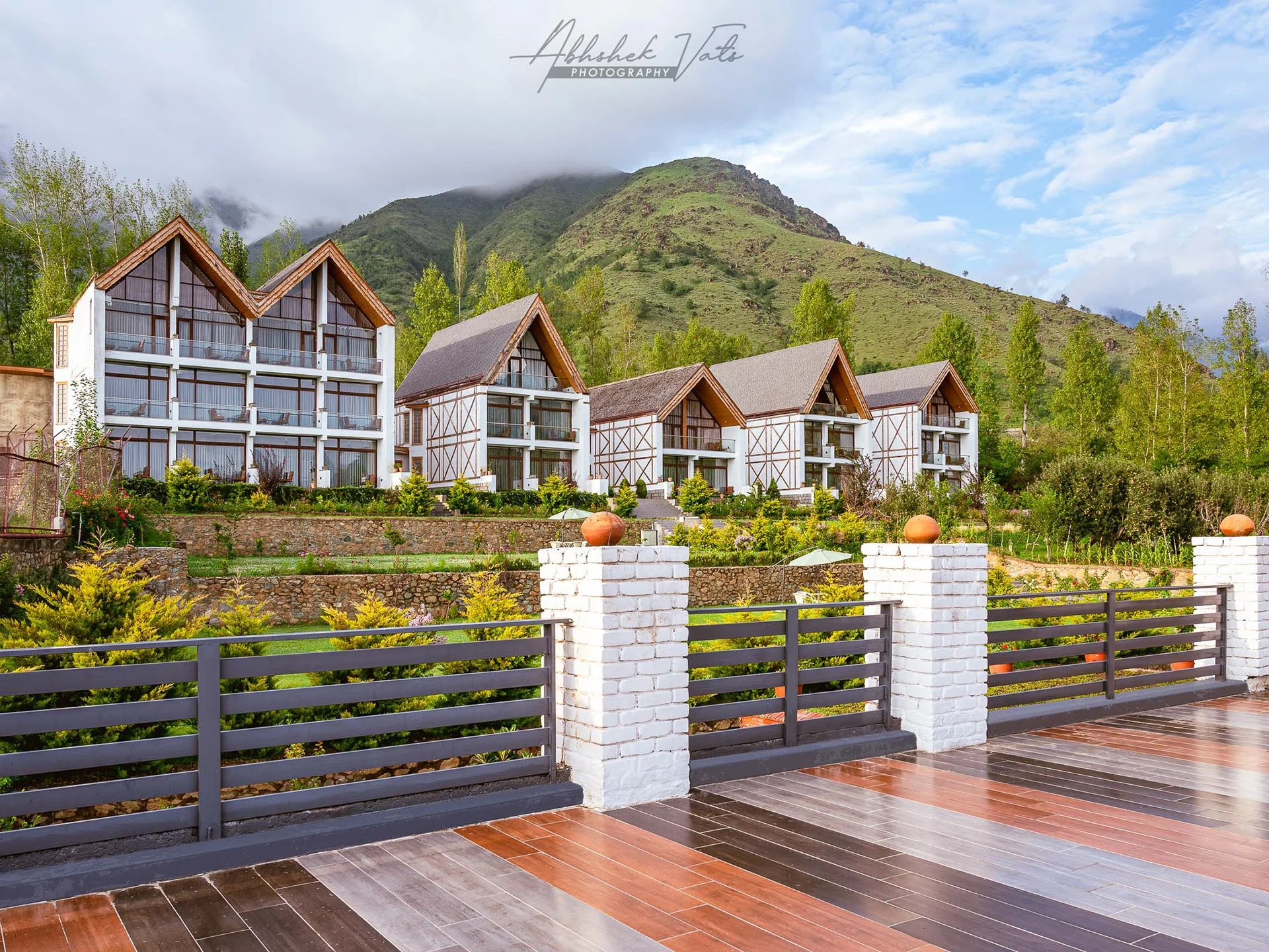 View of a modern residential complex with white buildings featuring wooden accents and glass balconies, surrounded by lush greenery and mountains in the background.