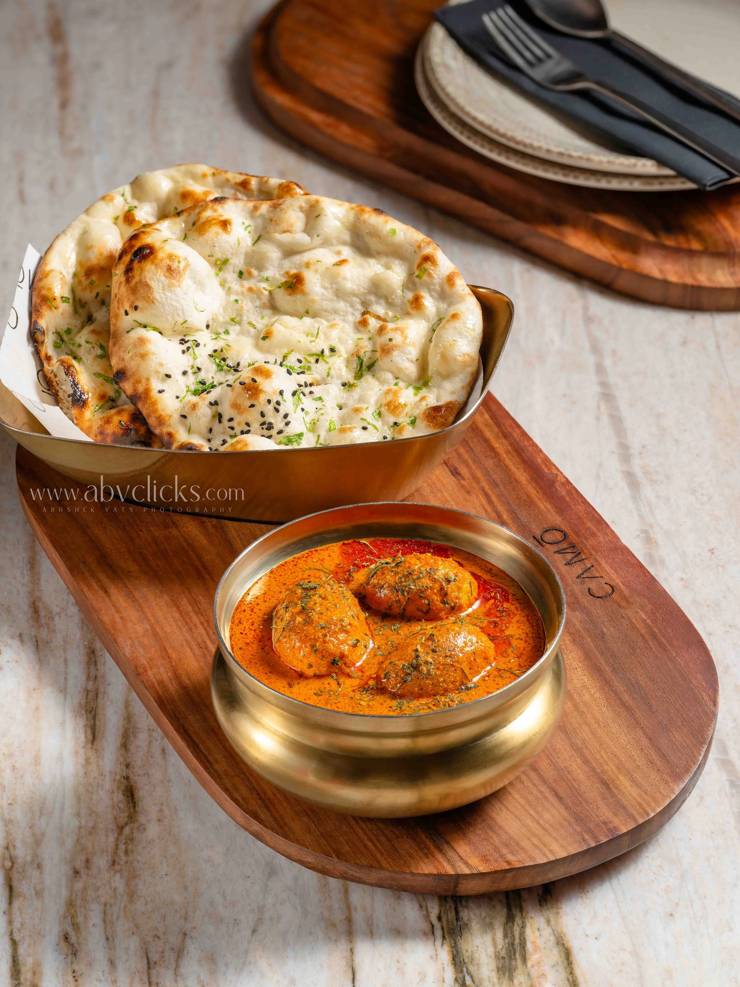 A wooden platter holding two dishes of Indian cuisine on a marble table. One is a bowl of naan bread garnished with herbs and black sesame seeds. The other is a bowl of curry with meatballs, coated in a red sauce with herbs.