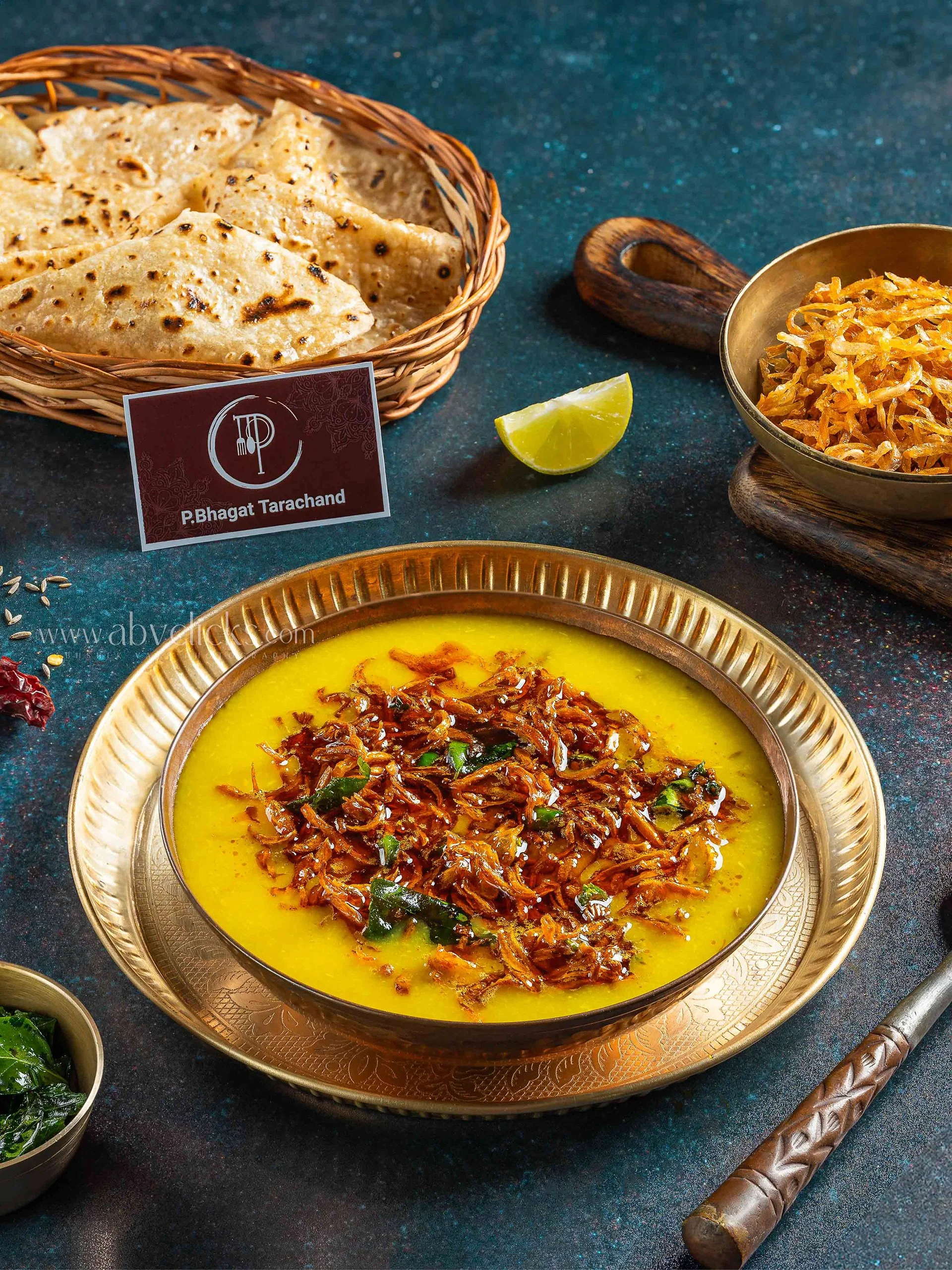 A traditional Indian dish called Dal Tadka served in a silver bowl with crispy papadums and a wedge of lemon on the side, placed on a black textured surface with a brass spoon and a decorative wooden handle.