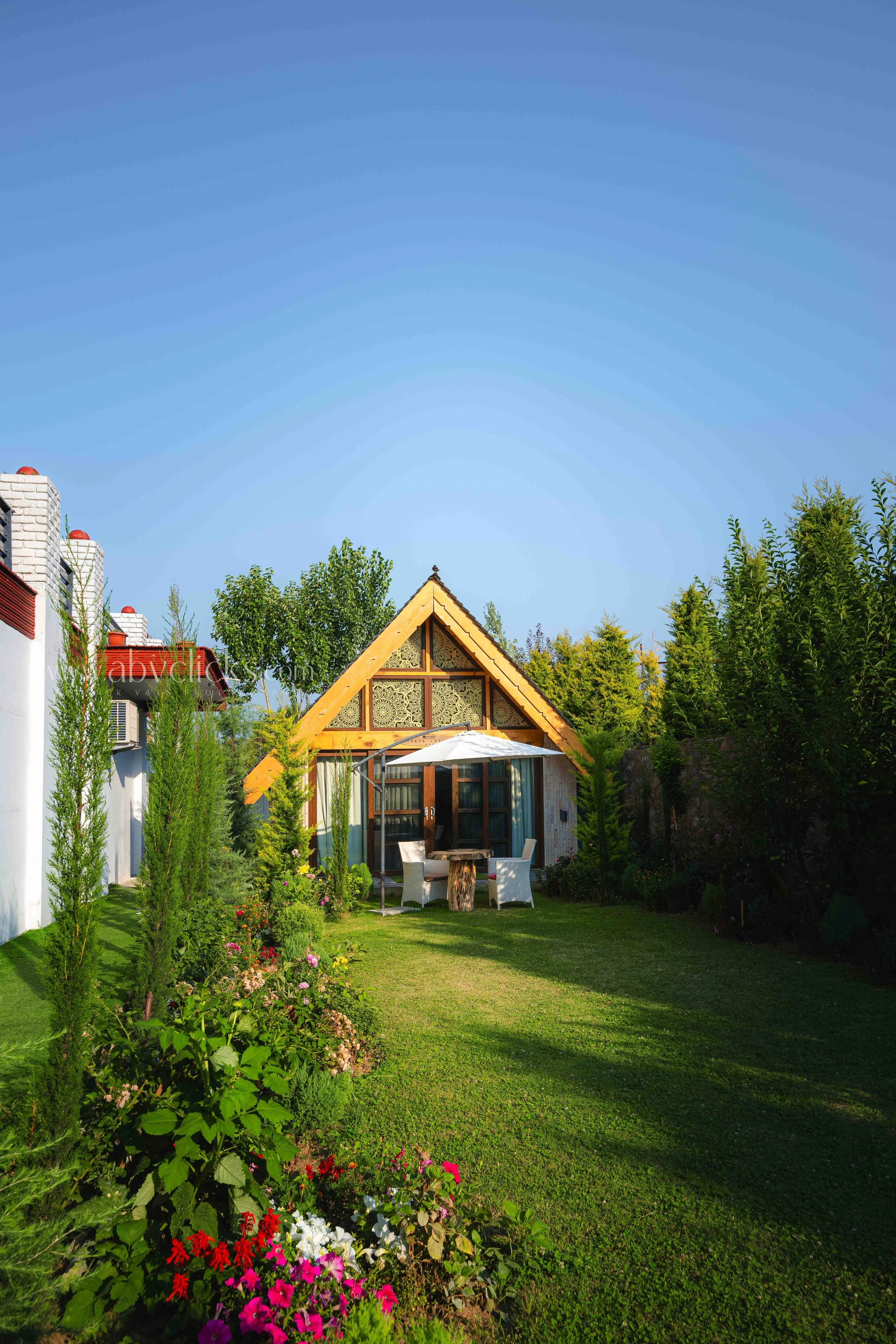 A lush green backyard garden with colorful flowers, a small wooden house with a triangular roof, and surrounded by tall trees under a clear blue sky.