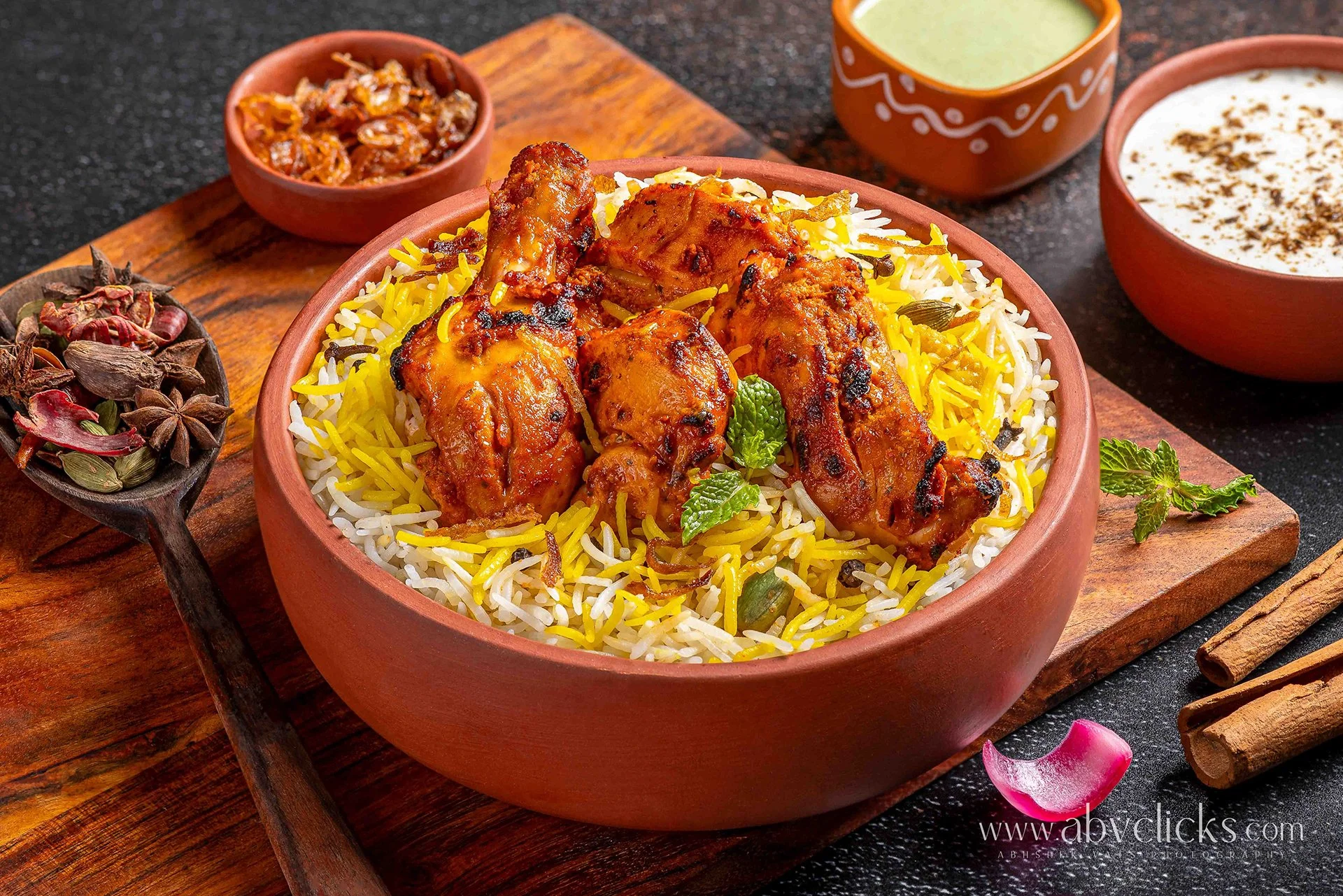 A wooden bowl filled with colorful saffron rice topped with grilled chicken drumsticks, garnished with mint leaves, on a wooden tray with bowls of raita, salad, and chutney in the background, set on a black surface.