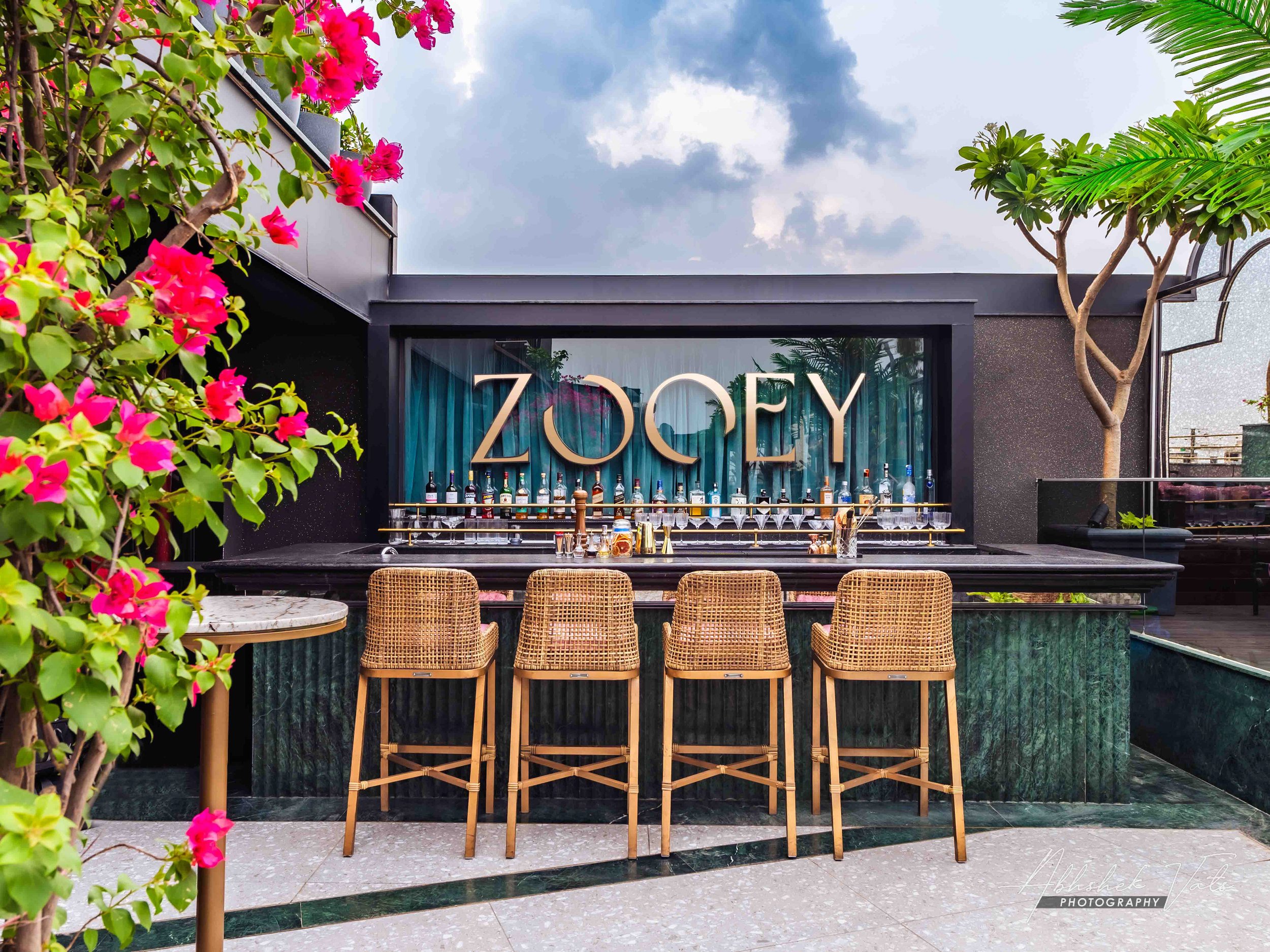 A modern outdoor bar with a black countertop and barstools, surrounded by lush green plants and pink flowers, with a cloudy sky in the background.