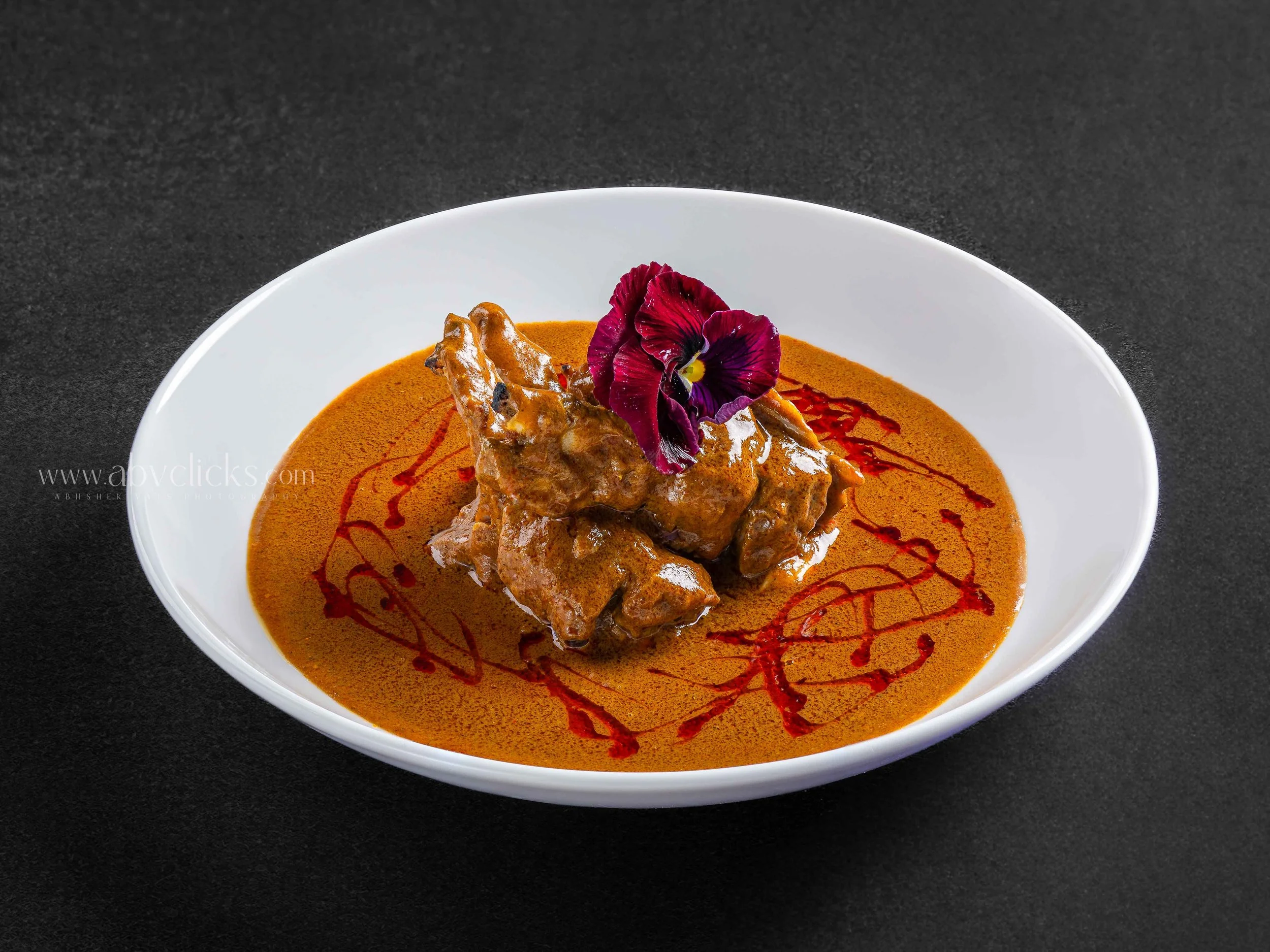 A white bowl of curry garnished with an edible flower and red sauce drizzle, placed on a black surface.