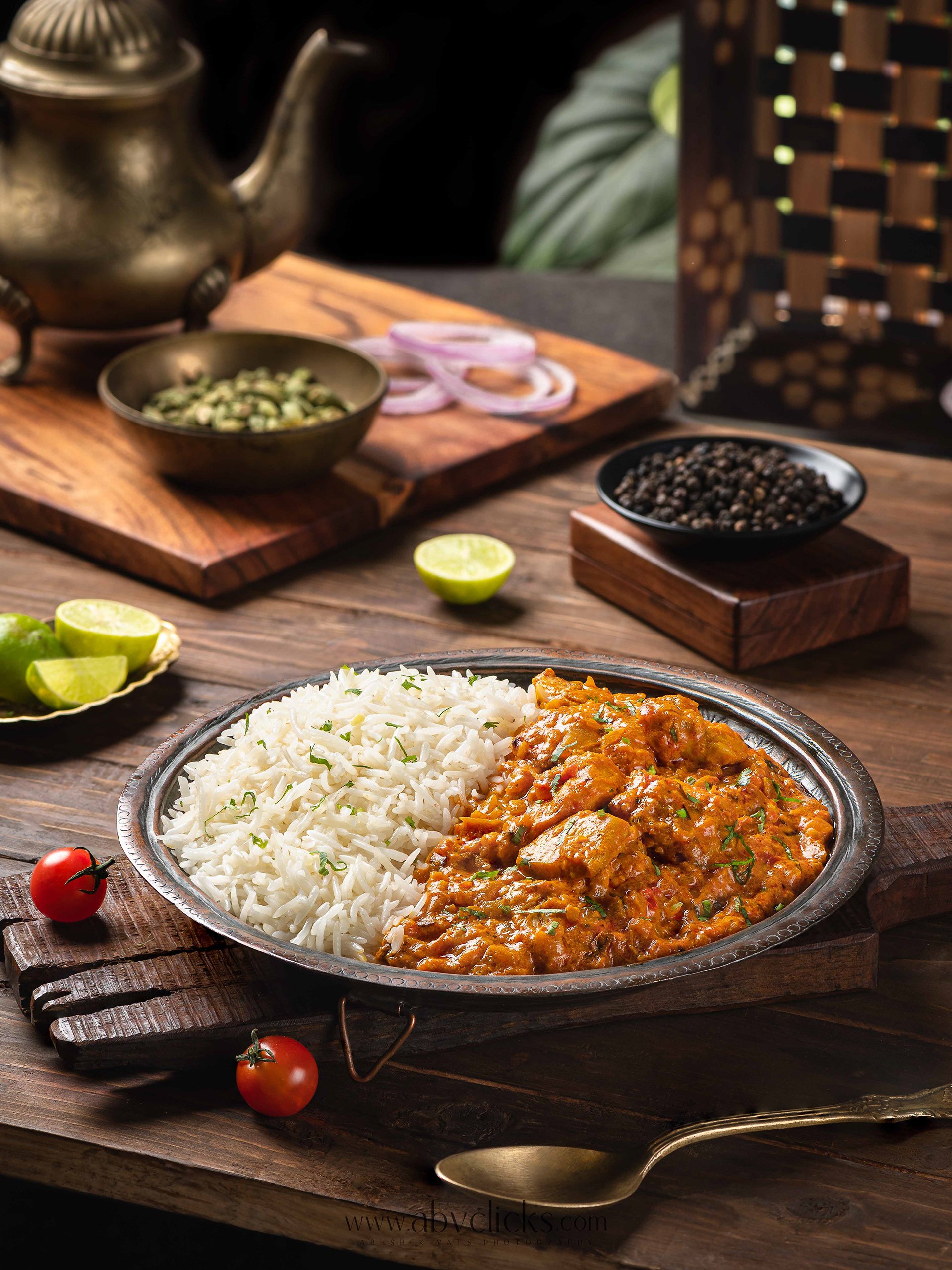 Plate of white rice and chicken curry with vegetables on a wooden table, with tomatoes and limes nearby, and various spices in the background.