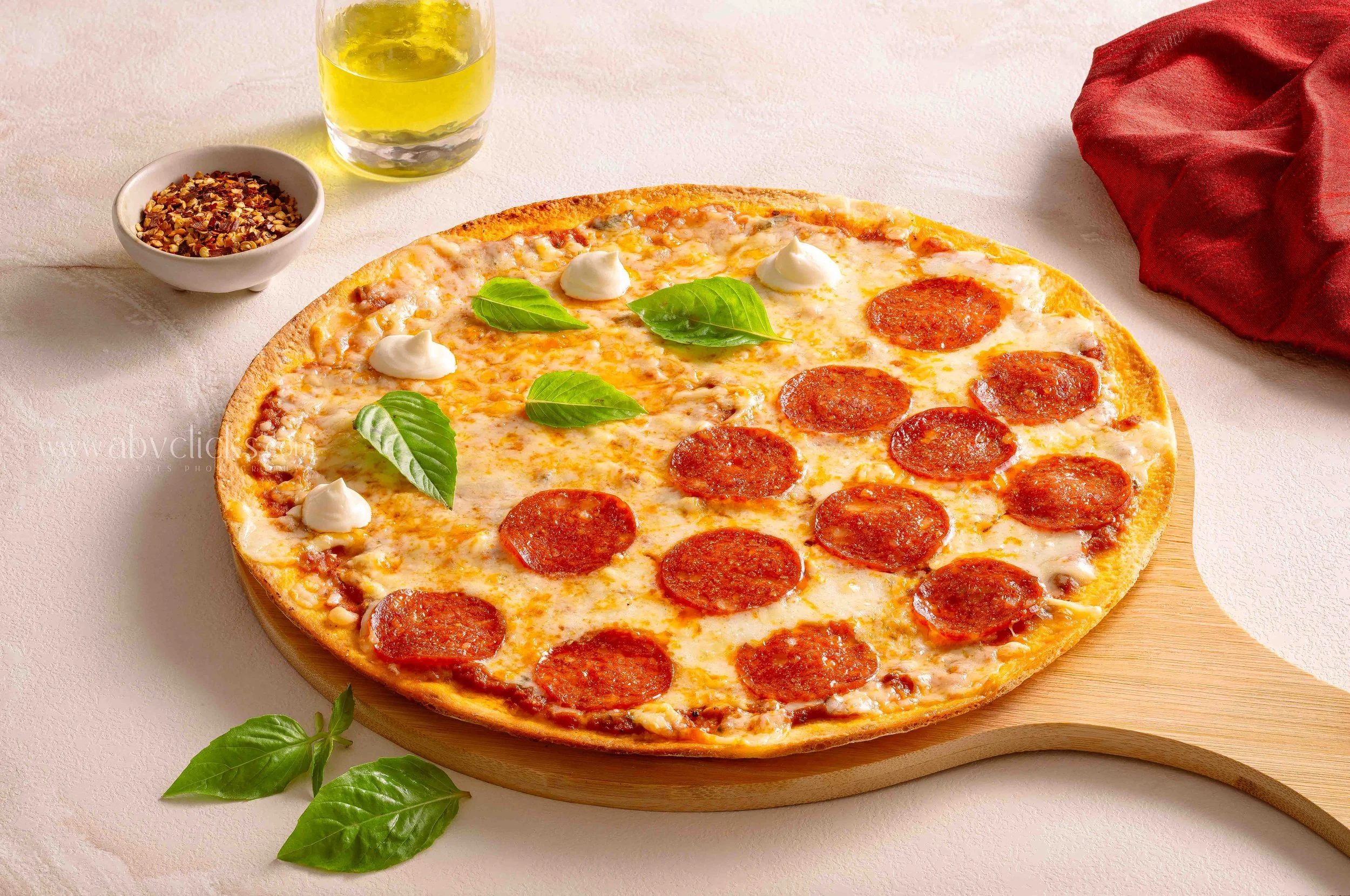 Cheese pizza with pepperoni slices on a round wooden board, garnished with fresh basil leaves, with a side of crushed red pepper flakes, a glass of yellow beverage, and a red cloth on a light-colored surface.