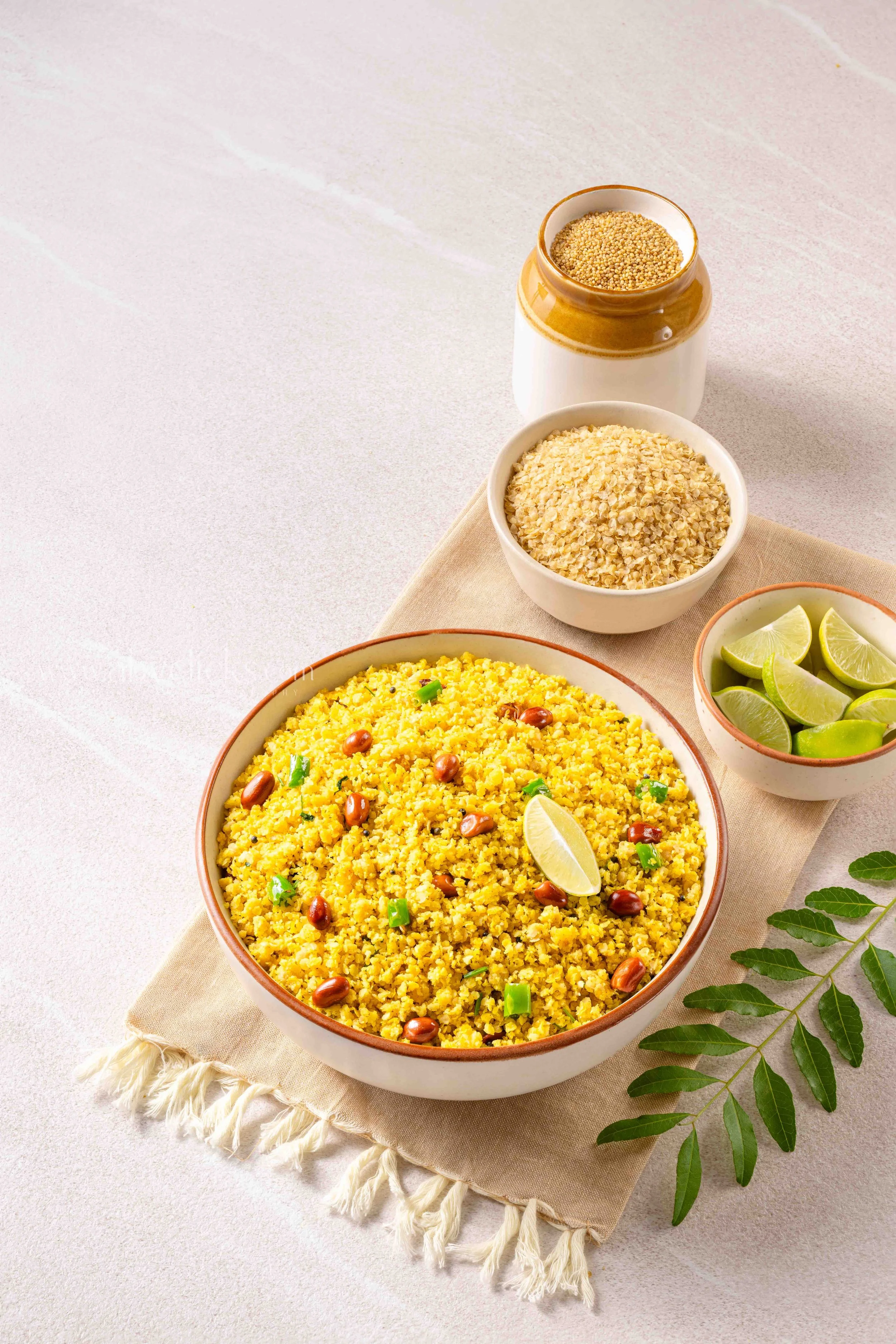 Bowl of yellow lentil dish garnished with peanuts, green chili, and a lemon wedge, with small bowls of millet, oats, and lime wedges nearby on a table.