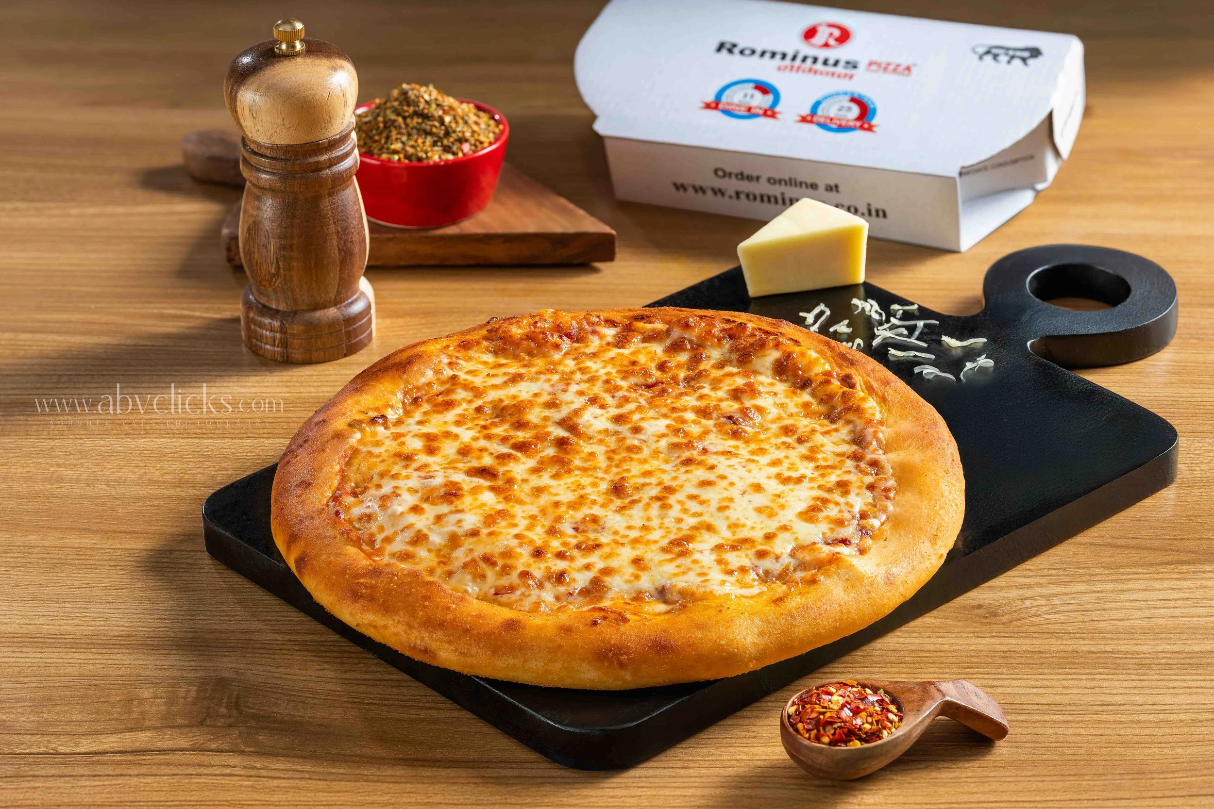 A cheese pizza on a black serving tray with cheese topping, a block of cheese, a container of chili flakes, a wooden pepper grinder, a container of ground spices on a wooden table, and a white pizza box in the background.