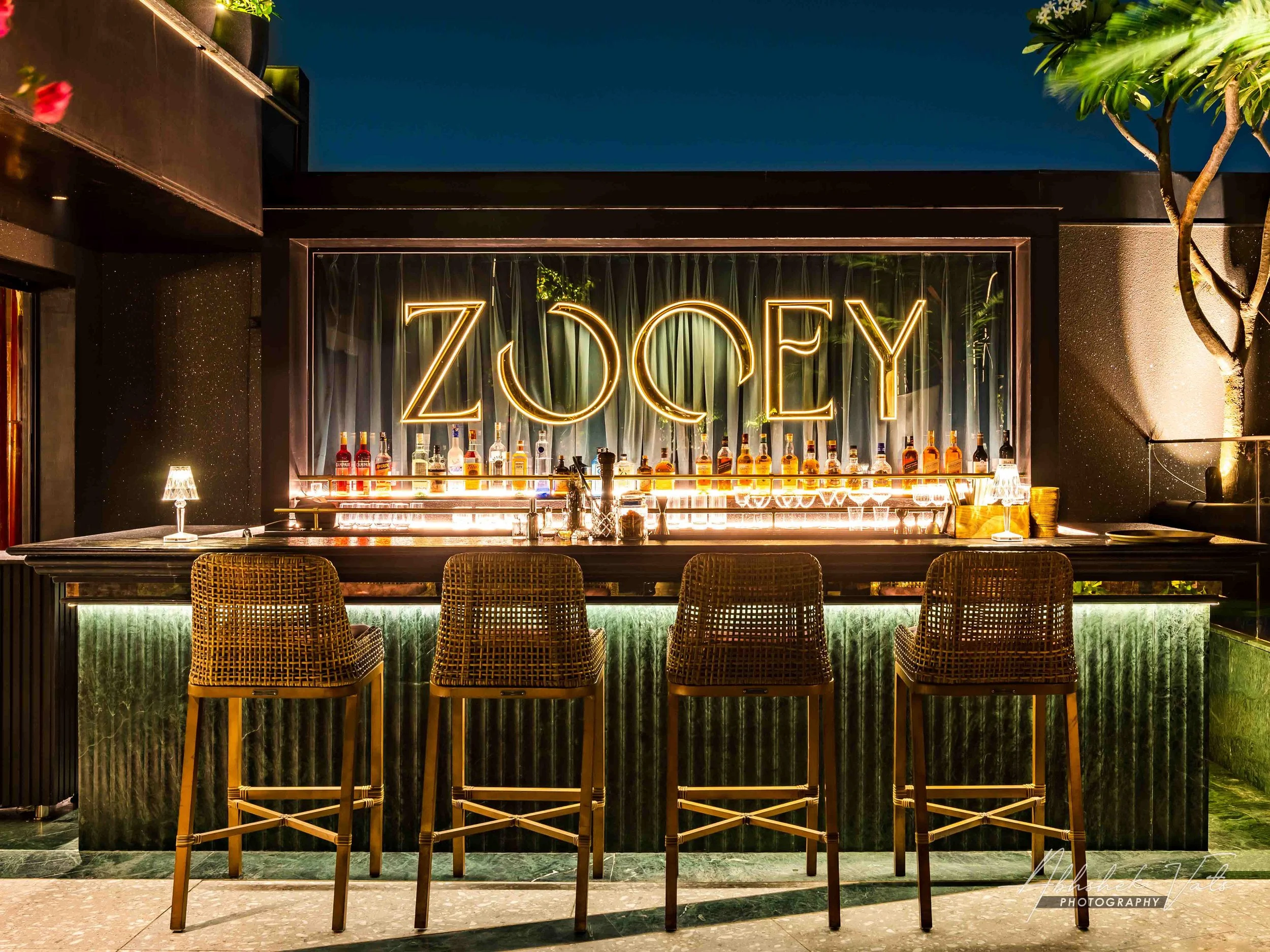 Bar area with a neon sign that reads 'ZOEY', bottles of liquor displayed behind the bar, woven chairs in front, and a tree to the right. Nighttime setting with warm lighting.