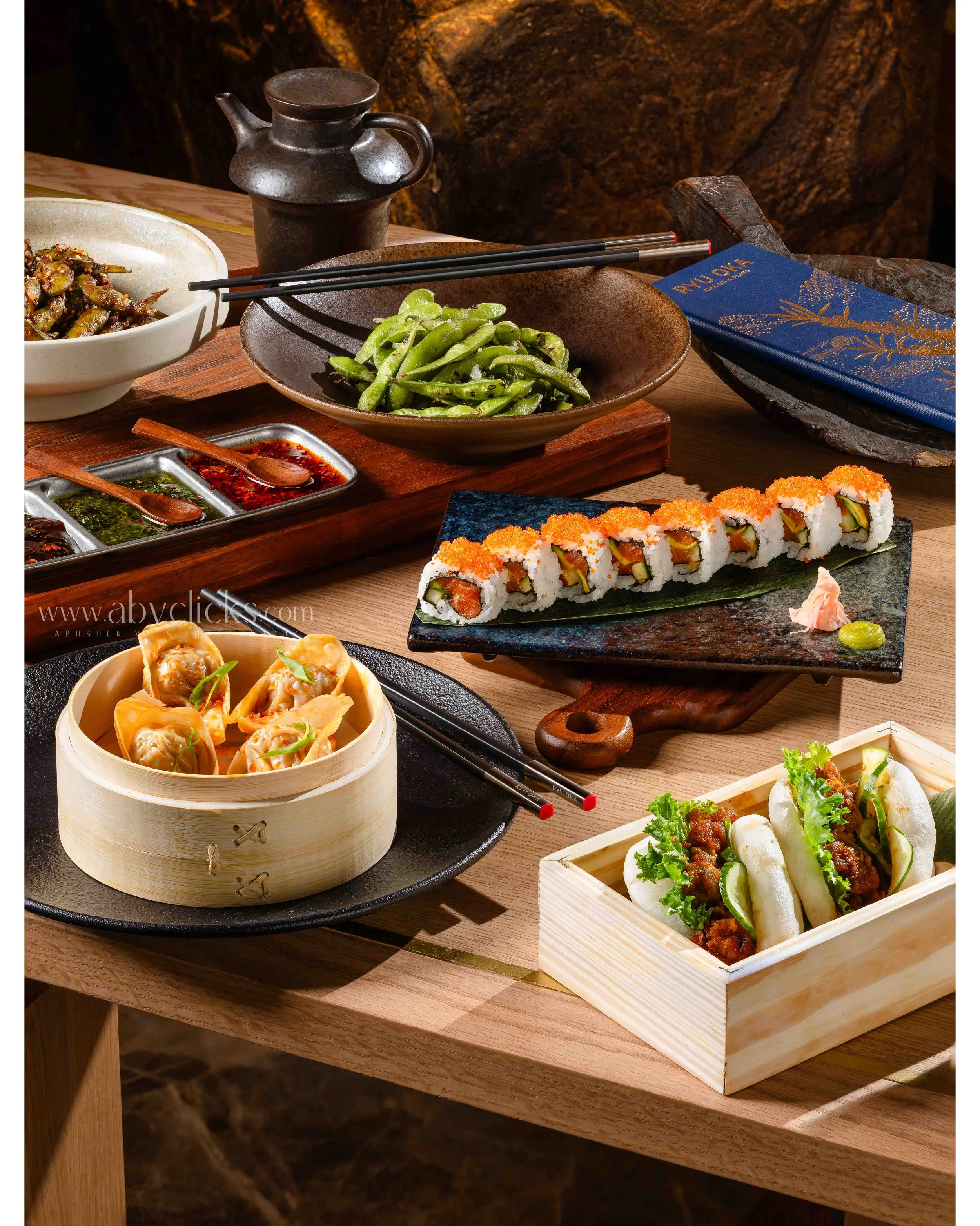 A variety of Japanese dishes displayed on a wooden table, including sushi rolls with orange roe and vegetables, steamed dumplings in a bamboo steamer, edamame beans in a brown bowl, and soy sauce with wooden spoons, with chopsticks resting on a dark 