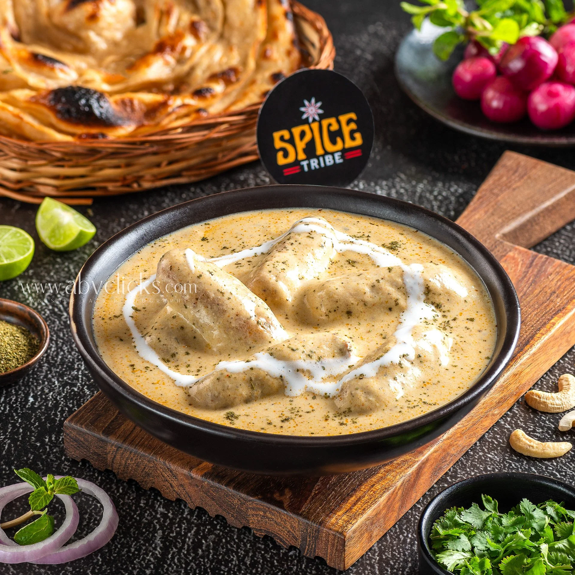 Bowl of chicken curry garnished with cream and herbs, surrounded by lime wedges, fresh herbs, sliced onions, and spices on a black countertop.