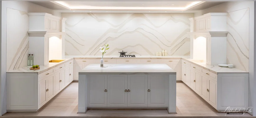 Minimalist white kitchen with custom cabinetry, marble backsplash, and a central island with a vase of flowers, under recessed lighting.