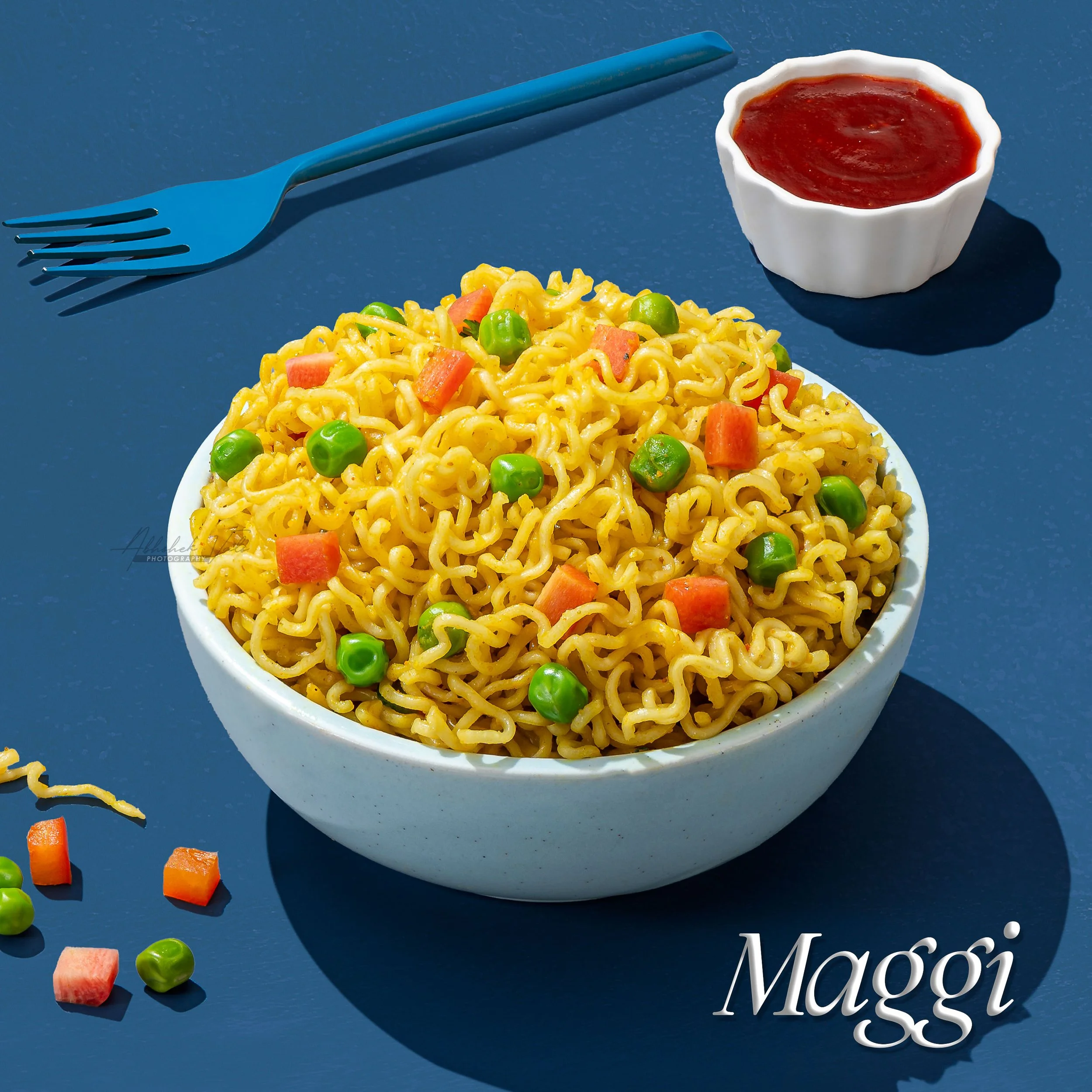 A bowl of cooked ramen noodles with peas and chopped carrots, a small white bowl of red sauce, and a blue fork on a dark blue surface.