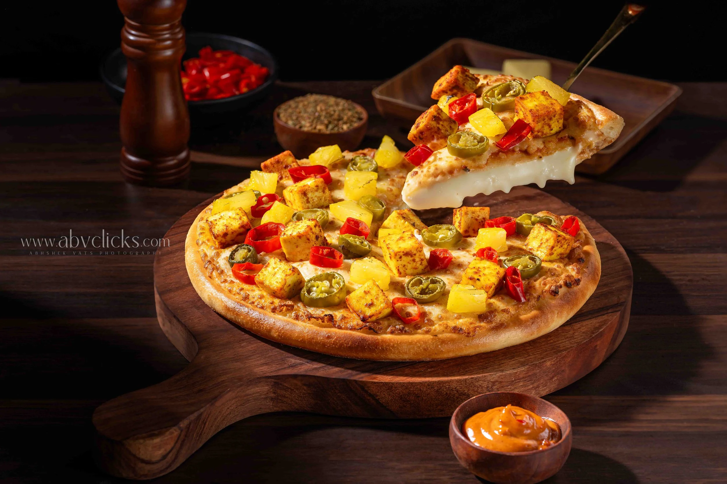 Close-up of a pizza topped with tofu cubes, red and green chili peppers, pineapple chunks, and melted cheese on a wooden serving board with small bowls of sliced chili peppers, seasonings, and dipping sauce in the background.