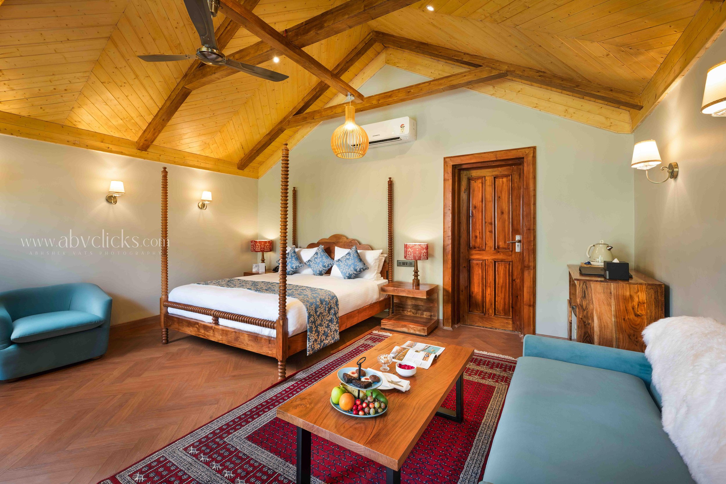 A cozy bedroom with a wooden vaulted ceiling, a four-poster bed with patterned pillows, a wooden door, a blue armchair, a wooden coffee table with snacks and fruit, wall-mounted lamps, and warm lighting.