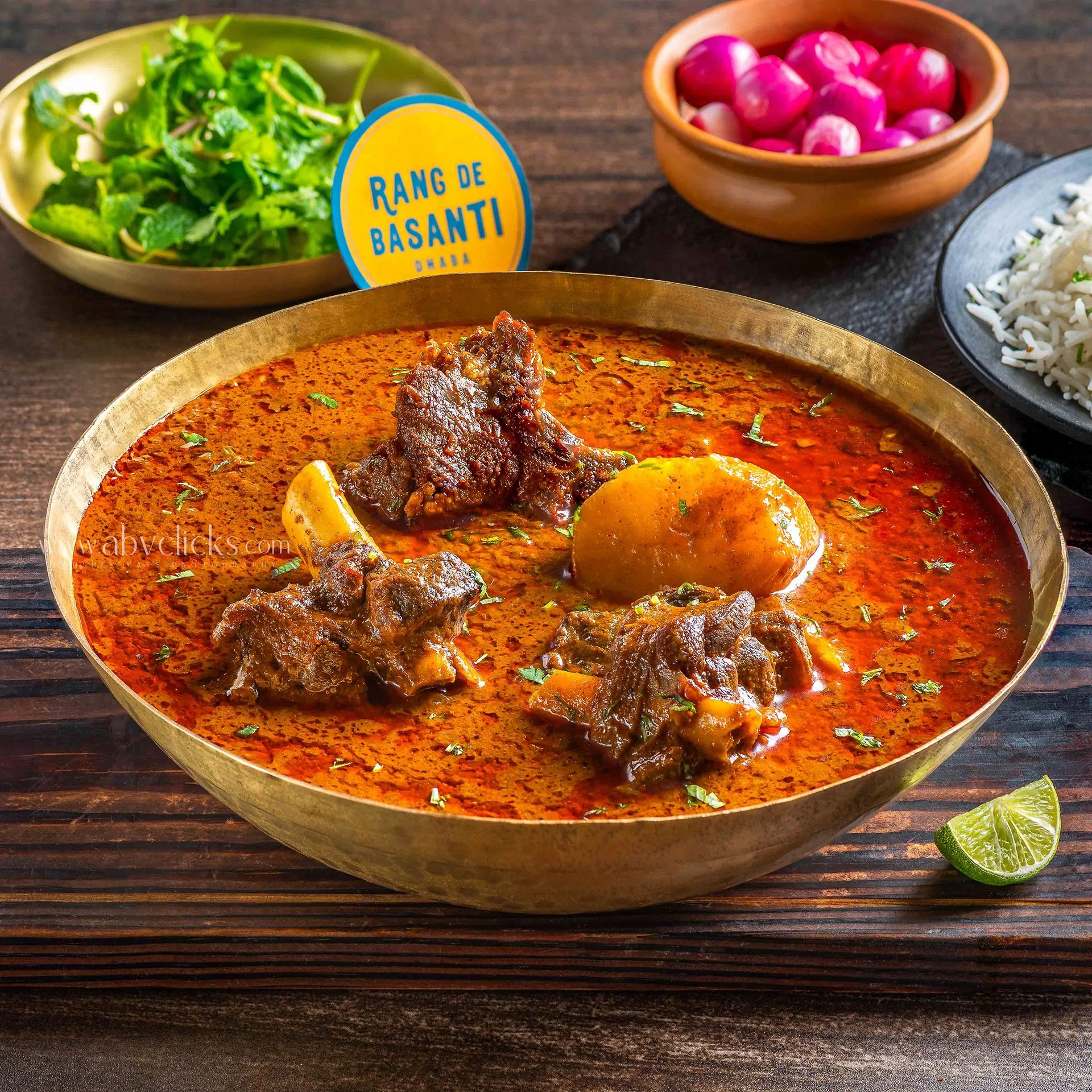A bowl of spicy Indian curry with beef, potatoes, and cilantro, accompanied by lime wedges, with side dishes of green salad, small pink onions, and white rice, on a dark wooden table.