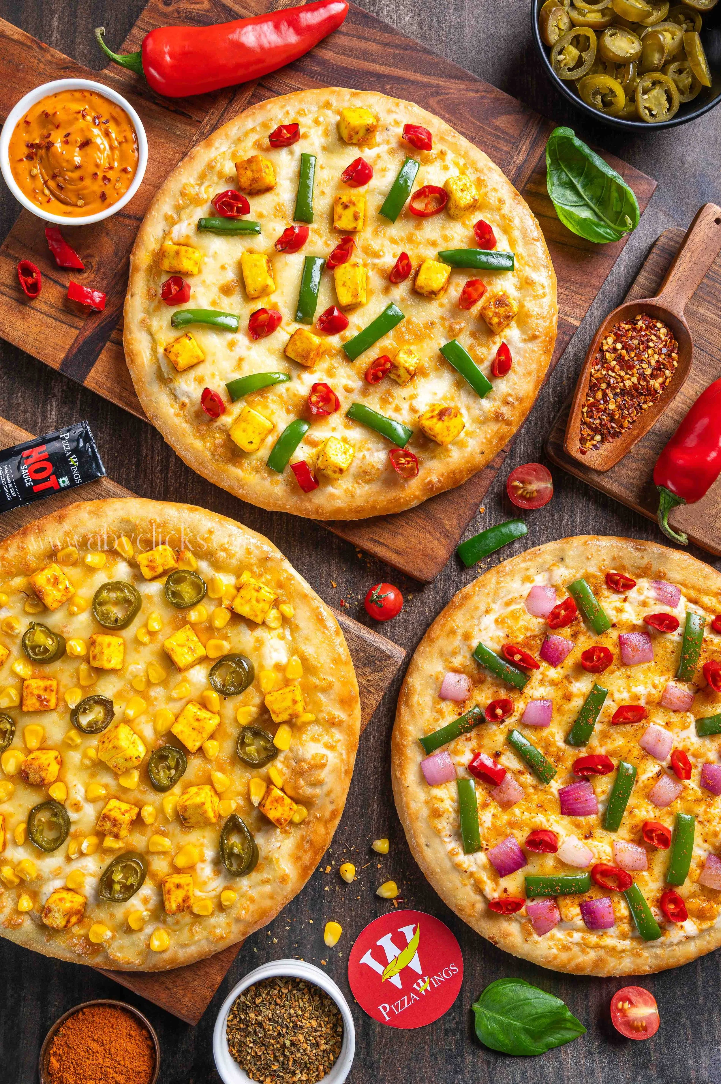 Three pizzas with various toppings such as jalapeños, corn, red chili peppers, green peppers, and diced chicken, set on a wooden board along with bowls of spices, sauces, and fresh herbs.