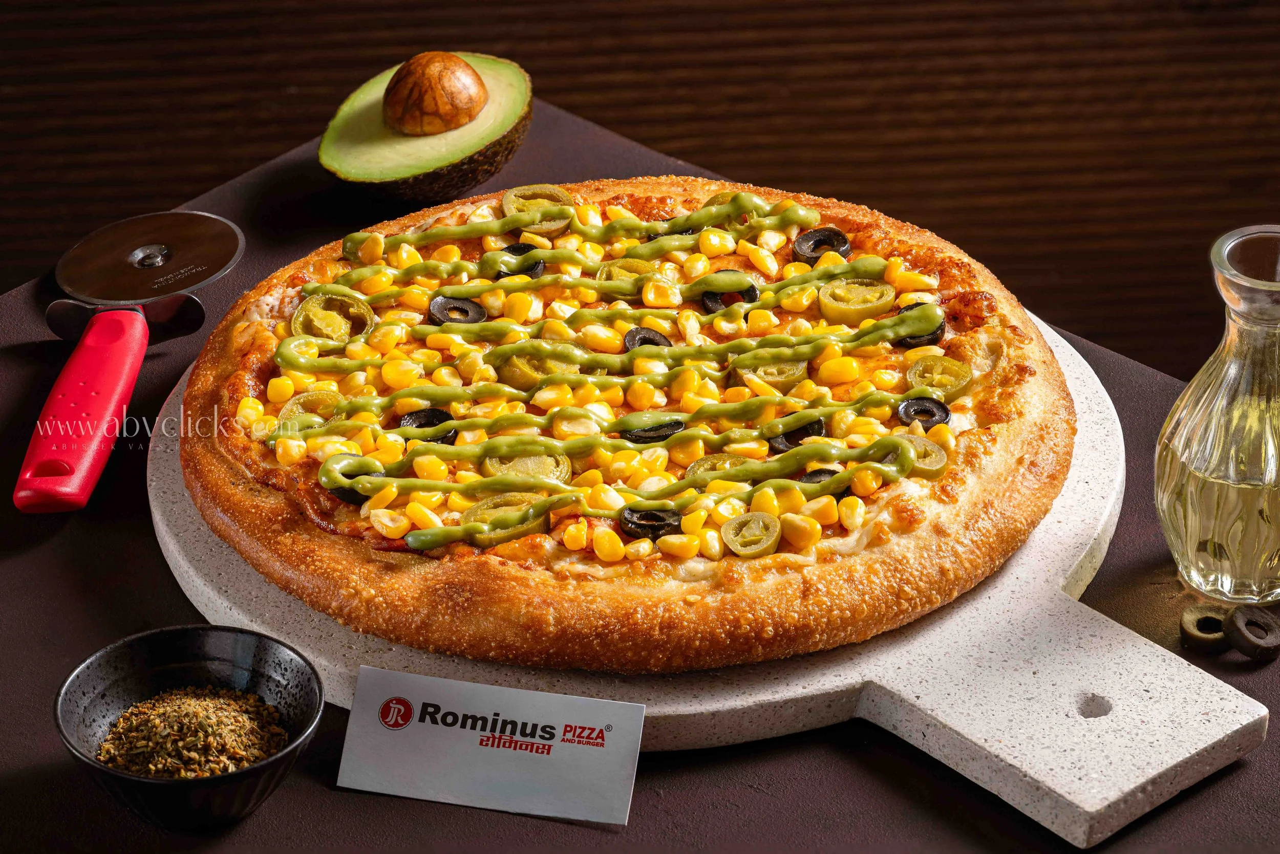 A large pizza topped with corn, black olives, and sliced green chilies on a white stone serving platter. A halved avocado with the pit still in one half is nearby, along with a small glass bottle of olive oil and a small bowl of spices. A pizza menu 