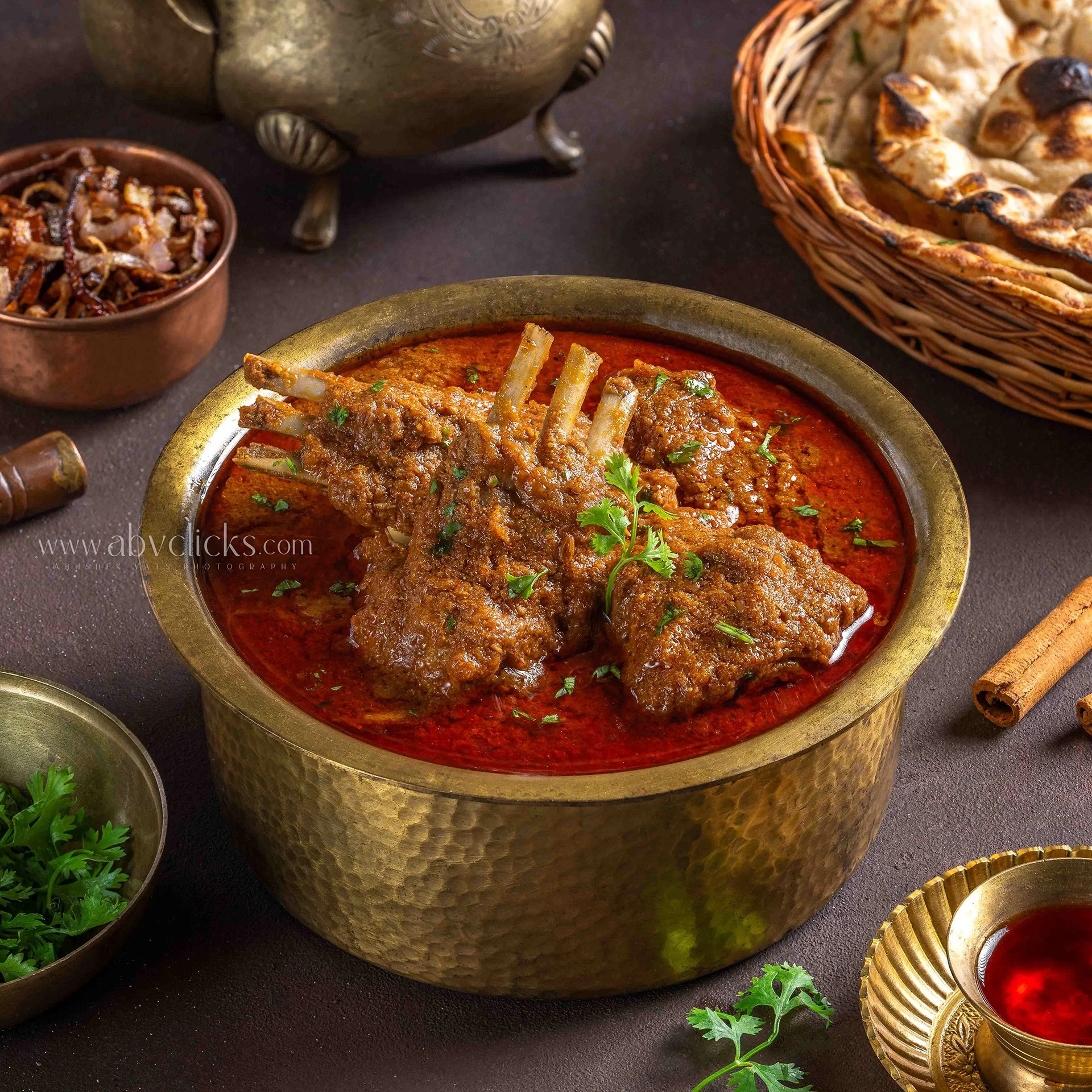 An Indian meal featuring a bowl of red lamb curry with bone-in meat garnished with cilantro, accompanied by naan bread in a woven basket, with small bowls of pickles and herbs nearby, set on a dark table.