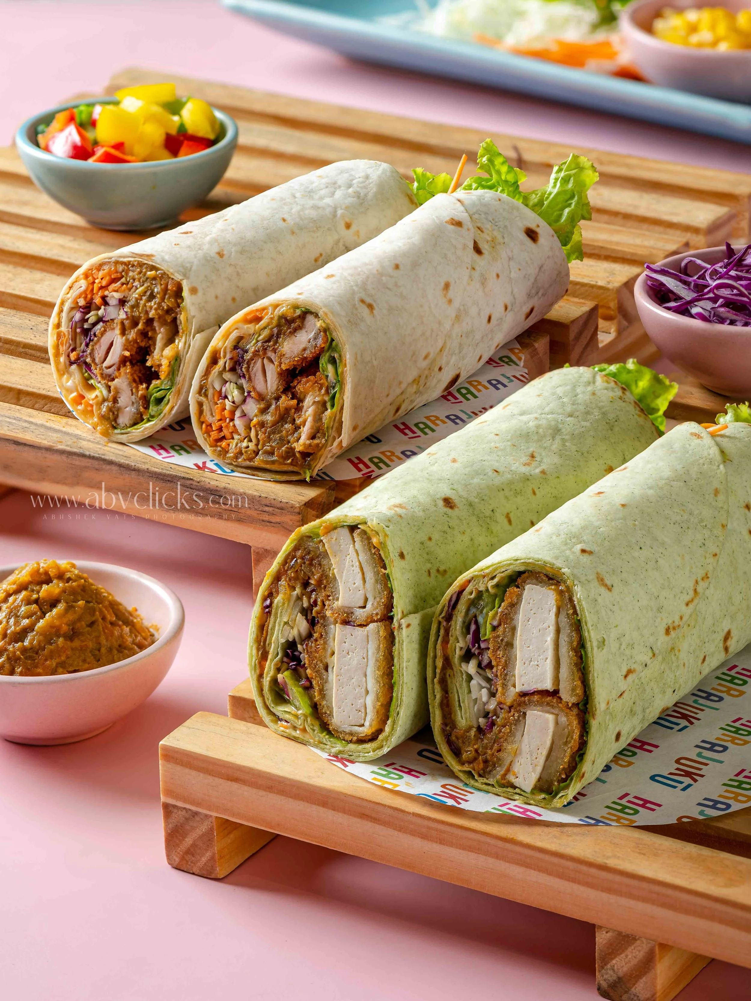 Two cut wraps with chicken, vegetables, and sauce on a wooden serving board, with side bowls containing salad, purple coleslaw, and a spicy sauce.