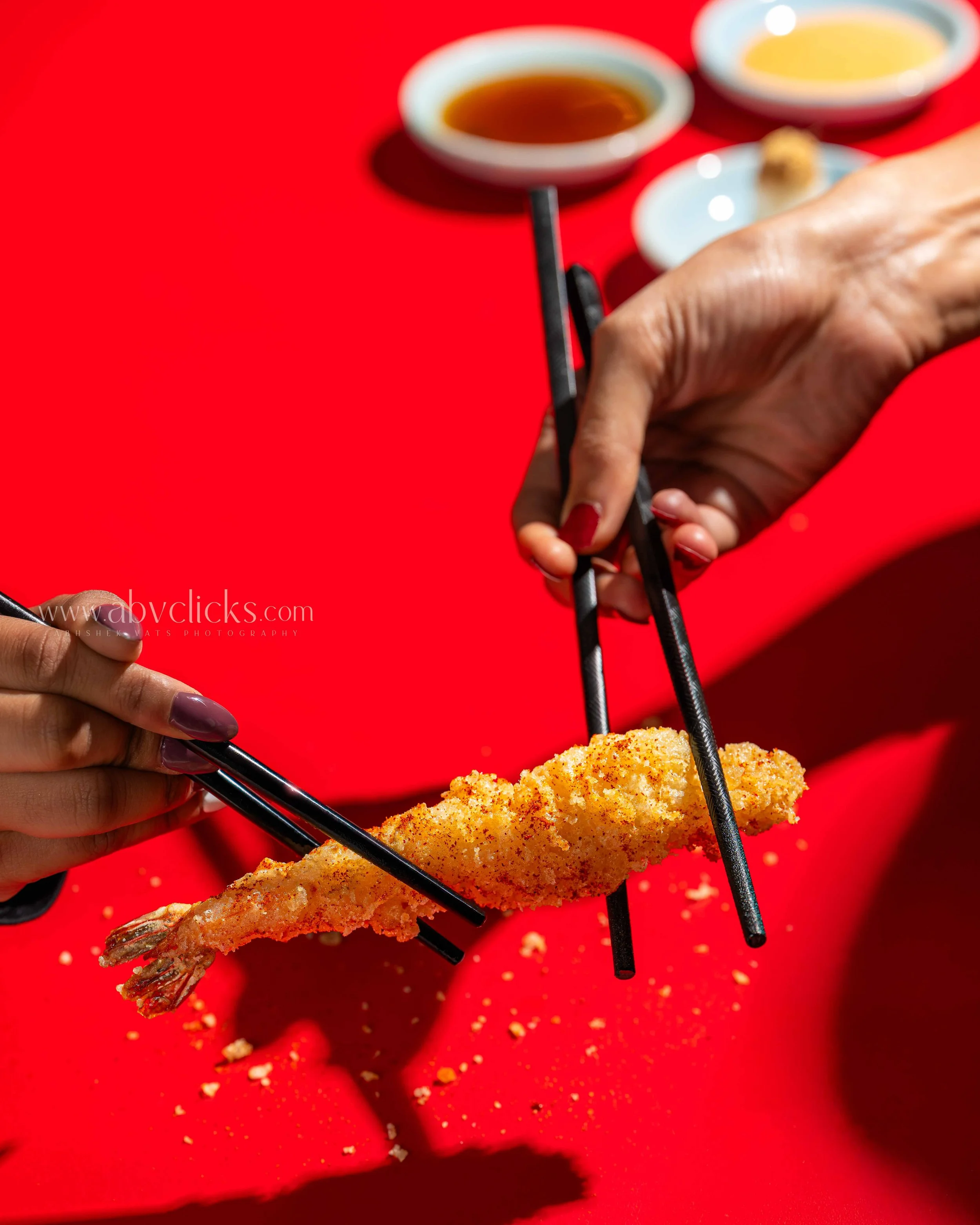 Close-up of a person's hand holding a pair of chopsticks, picking up a piece of breaded fried shrimp with a red background and small dishes of dipping sauces in the background.