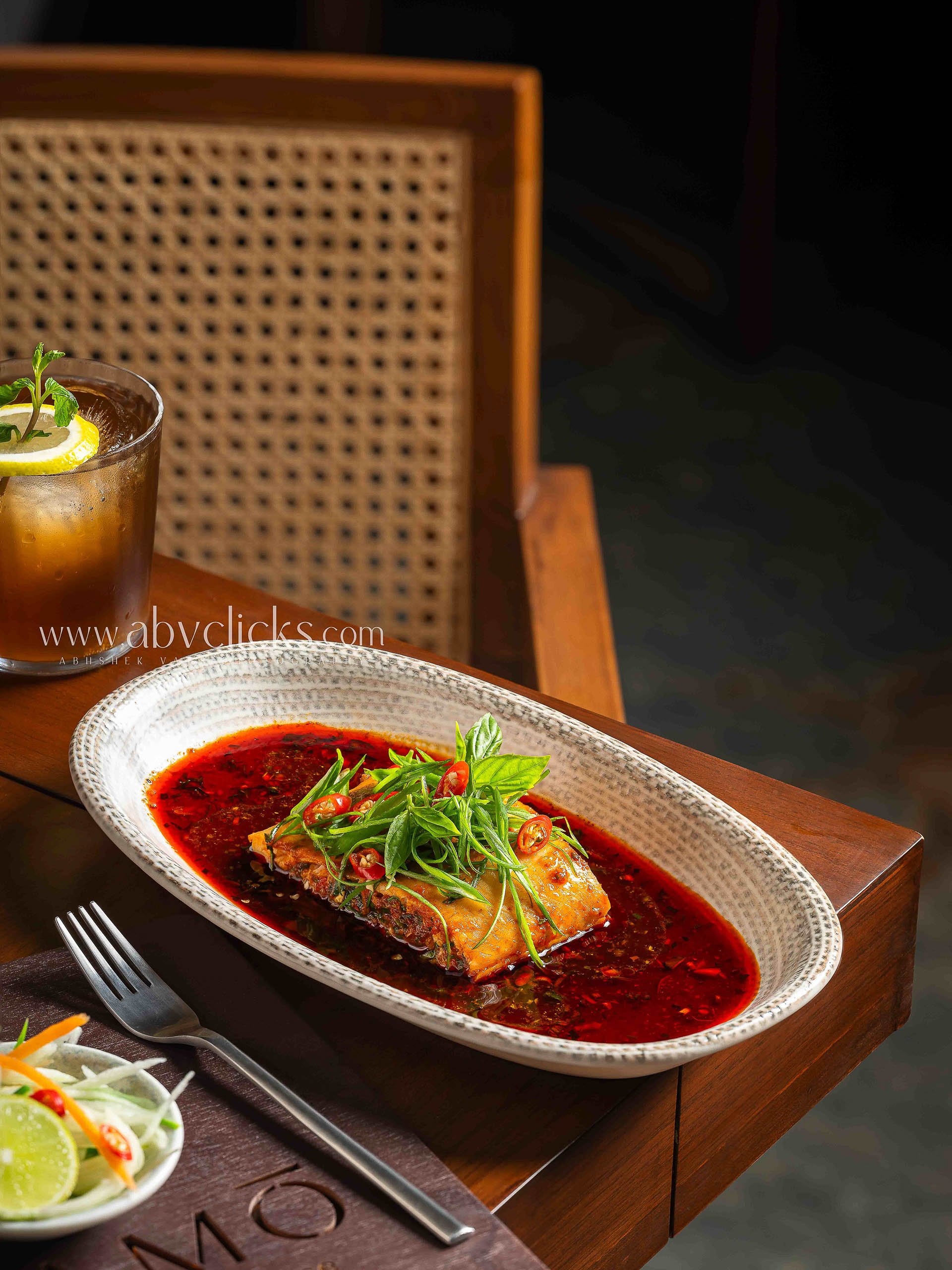 A fish dish served in a white oval bowl with a red sauce, garnished with green herbs and sliced red chilies, placed on a wooden table. A glass of iced tea with lemon and mint leaves is in the background.