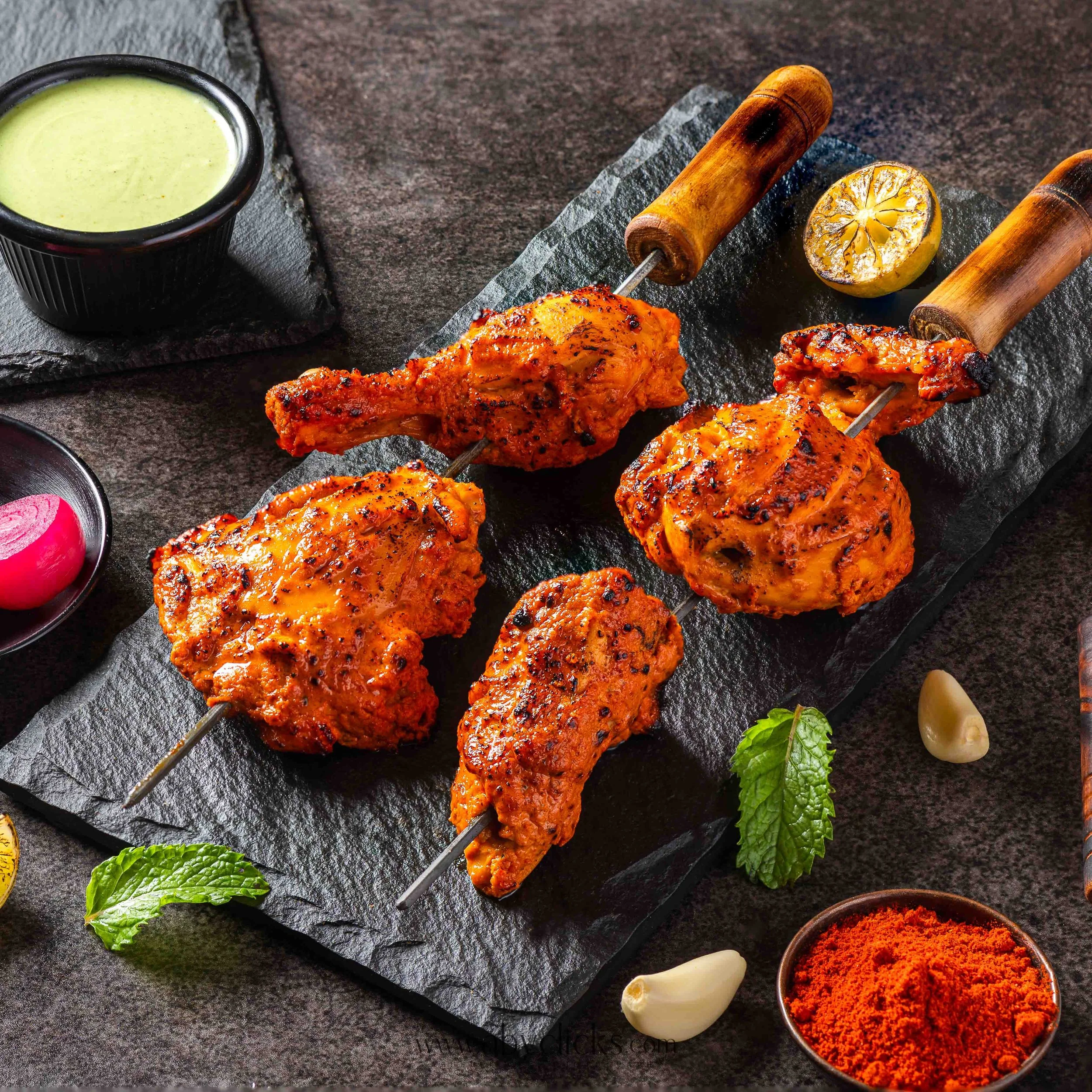 Skewers of grilled chicken in spicy marinade on black slate platter with lemon half, garlic, mint leaves, and small bowl of red spice powder.