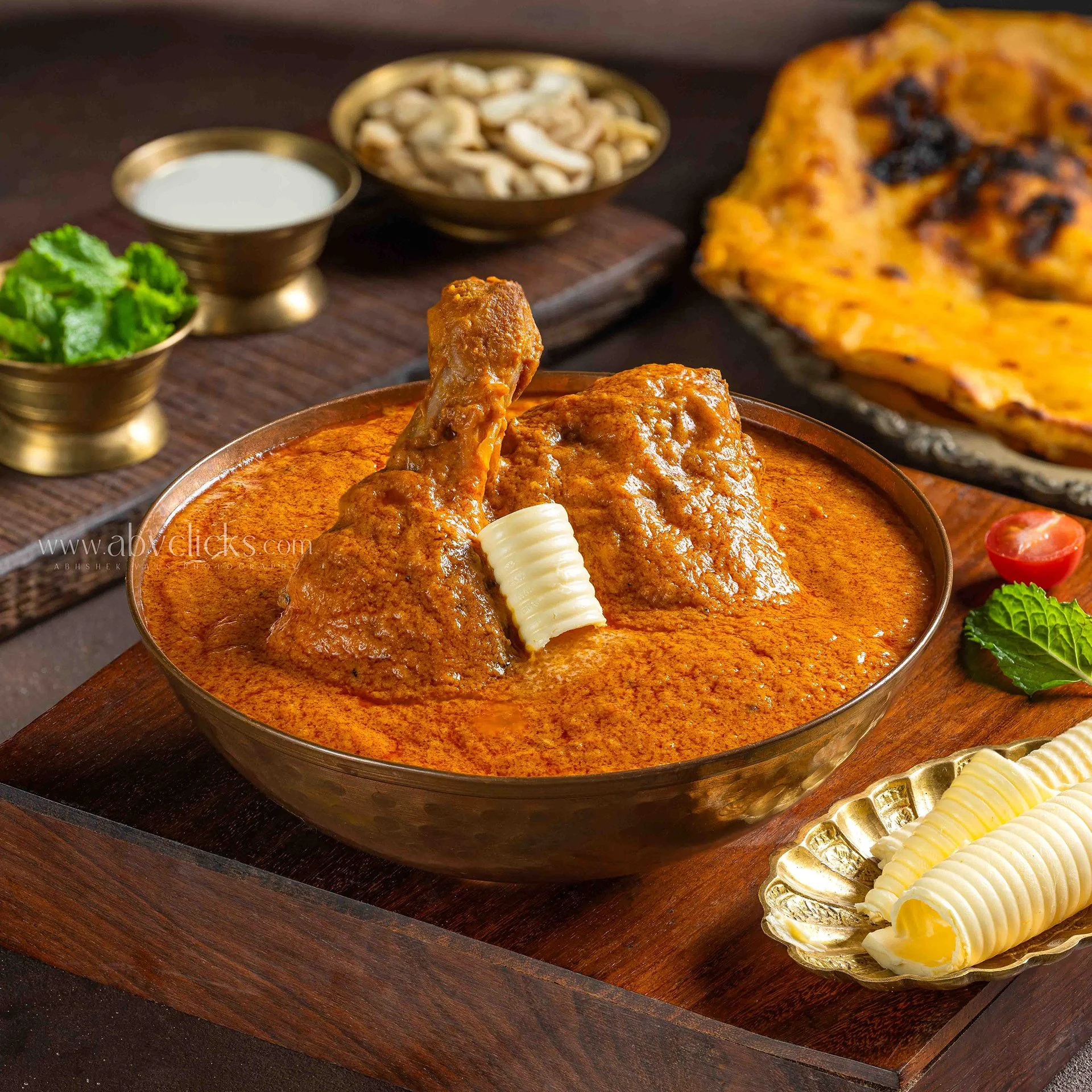 Close-up of Indian chicken curry with rice, garnished with butter and served on a wooden platter with side dishes including chopped greens, cashews, and a small bowl of white sauce.