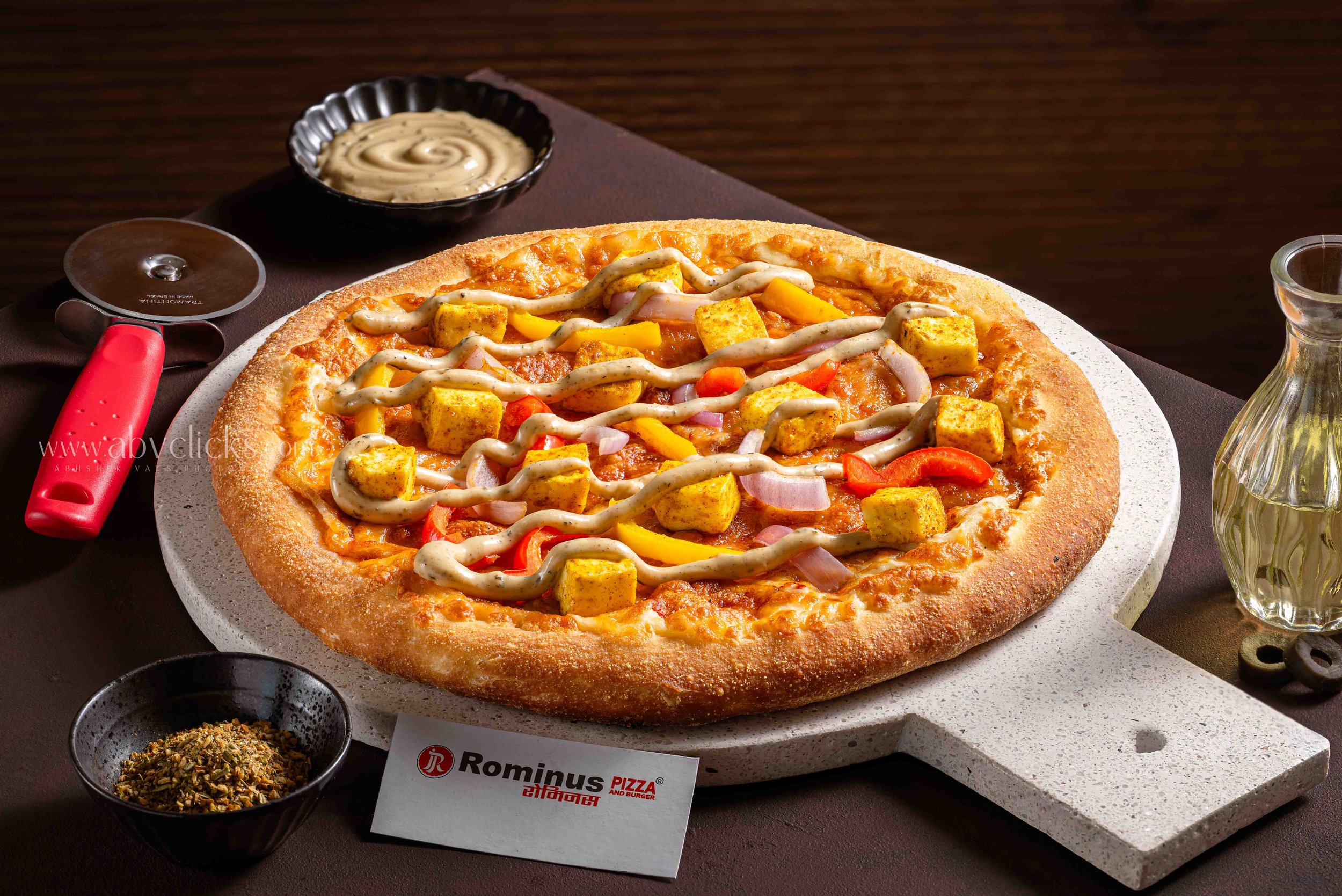 A pizza with various toppings including cheese, sausage, peppers, and onions on a round stone slab, with a small bowl of herb seasoning, a small bowl of sauce, a pizza cutter, and a glass of water nearby.