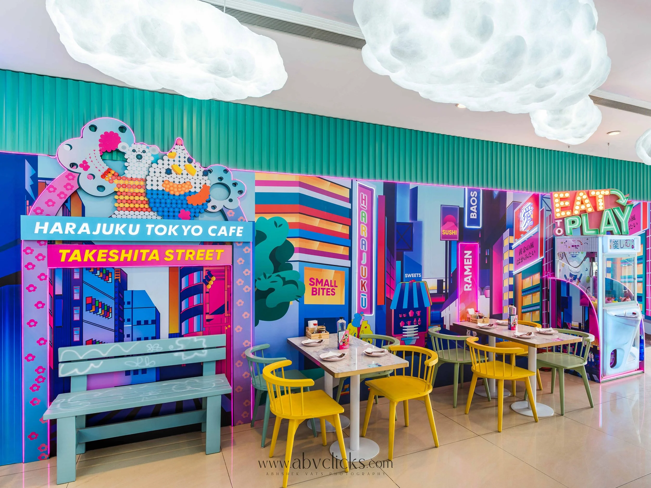 Colorful indoor cafe decorated with neon signs, cloud-shaped ceiling lights, and illustrated cityscape walls; features yellow and teal chairs with a white table, and a window with a claw machine in the background.