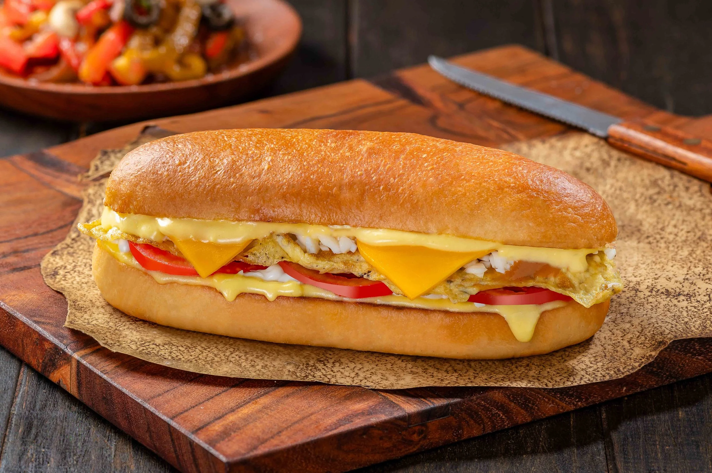 A submarine sandwich with slices of tomato, cheddar cheese, and egg, served on a wooden cutting board.