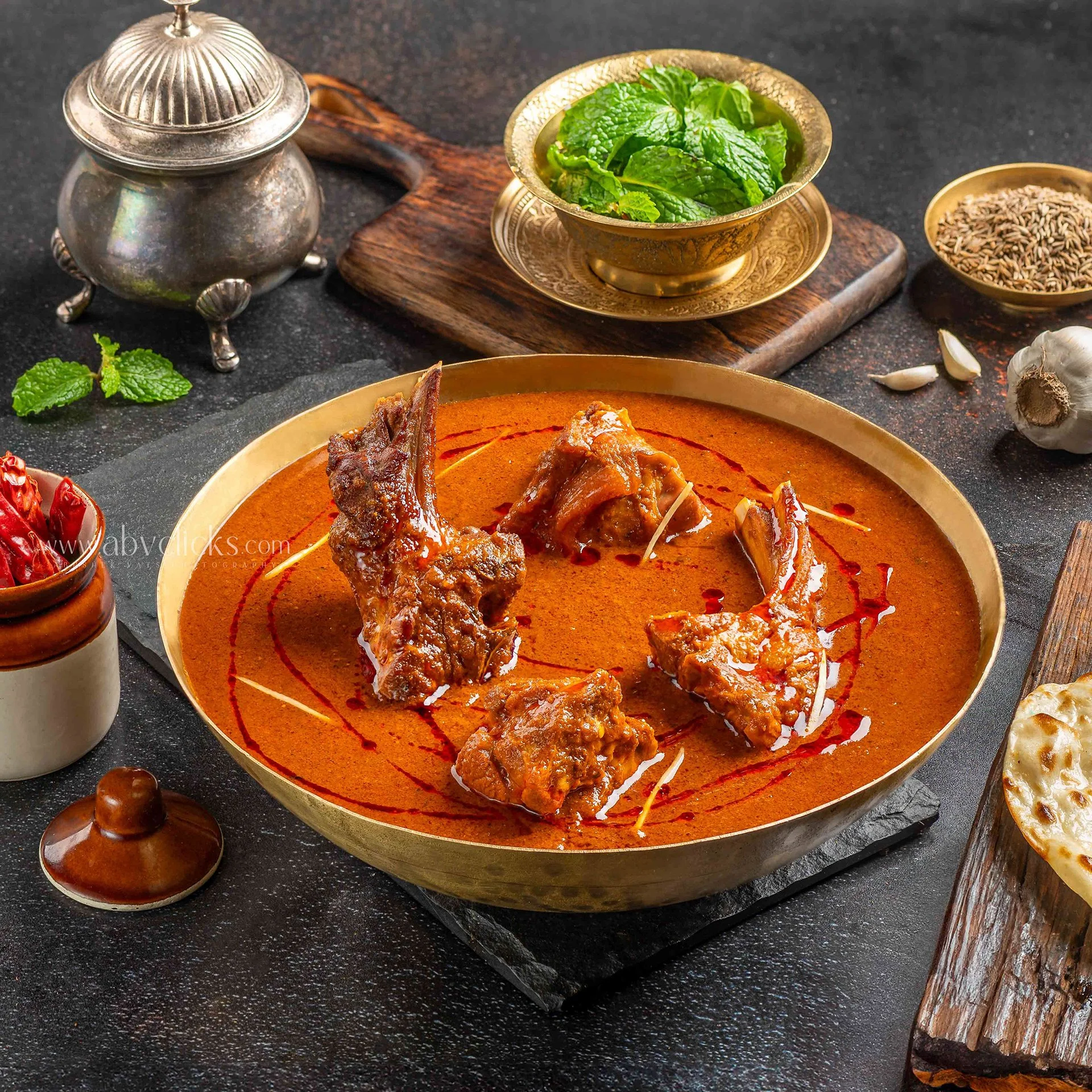 Traditional Indian dish, possibly curry, served in a golden bowl with meat pieces on top. Surrounded by small bowls of spices, garlic, and herbs on a dark table surface, with decorative elements.