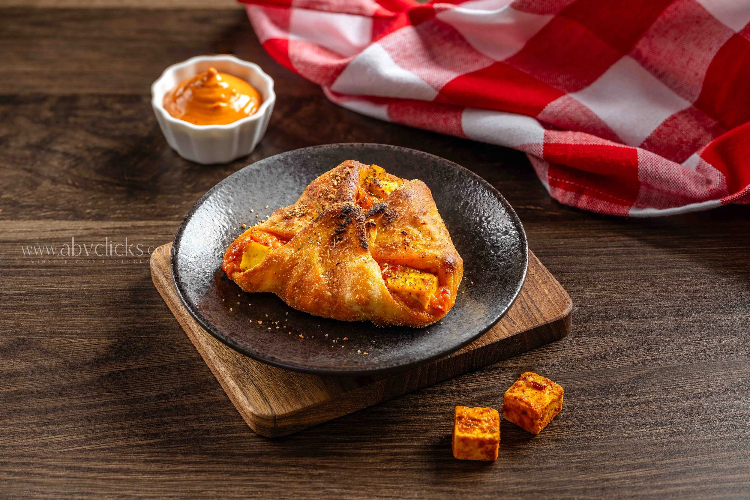 A slice of baked pizza with pepperoni and cheese on a round black plate, garnished with herbs, on a wooden serving board, accompanied by a small white bowl of sauce and a red and white checkered napkin on a wooden surface.