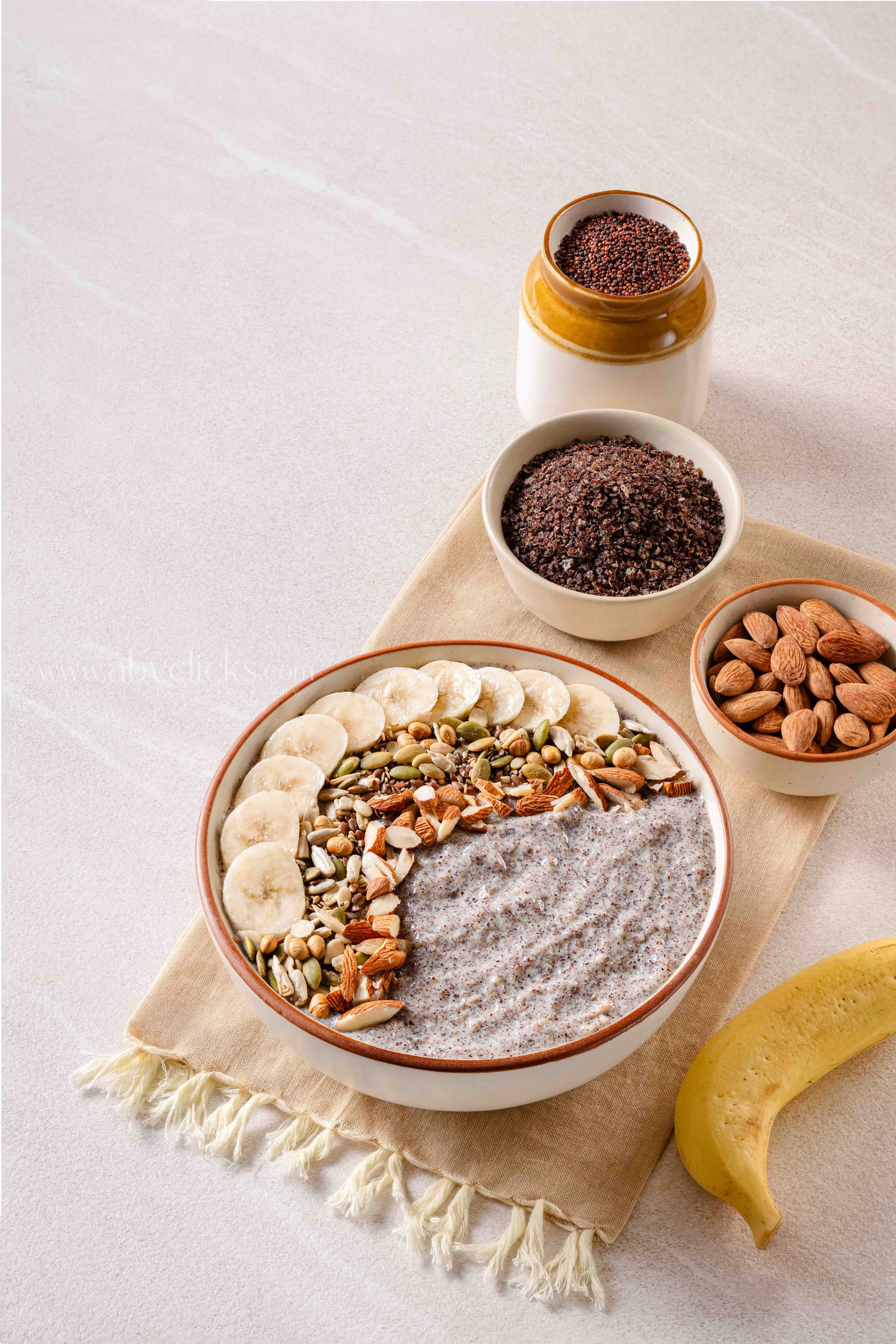 A bowl of chia seed pudding topped with banana slices, assorted seeds, and nuts. Next to the bowl are a small bowl of almonds, a jar of chia seeds, and a banana on a beige cloth.
