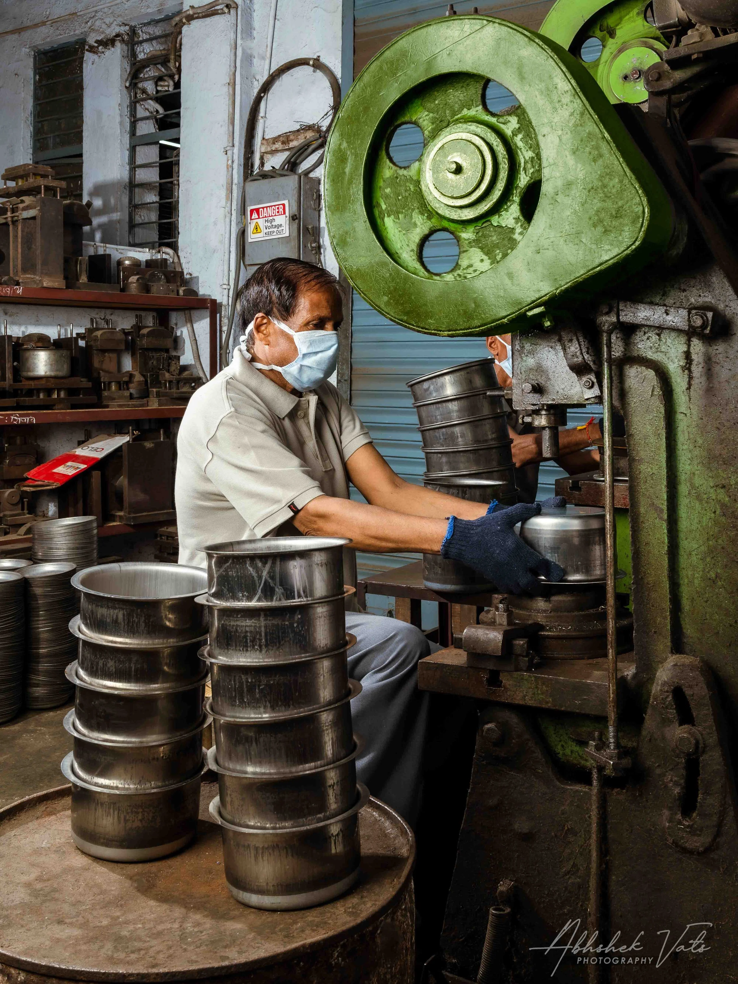 A man wearing a face mask working with metal components near a large, green industrial machine in a workshop. He is handling metal cylindrical objects, and there are stacked similar objects on the table in front of him.