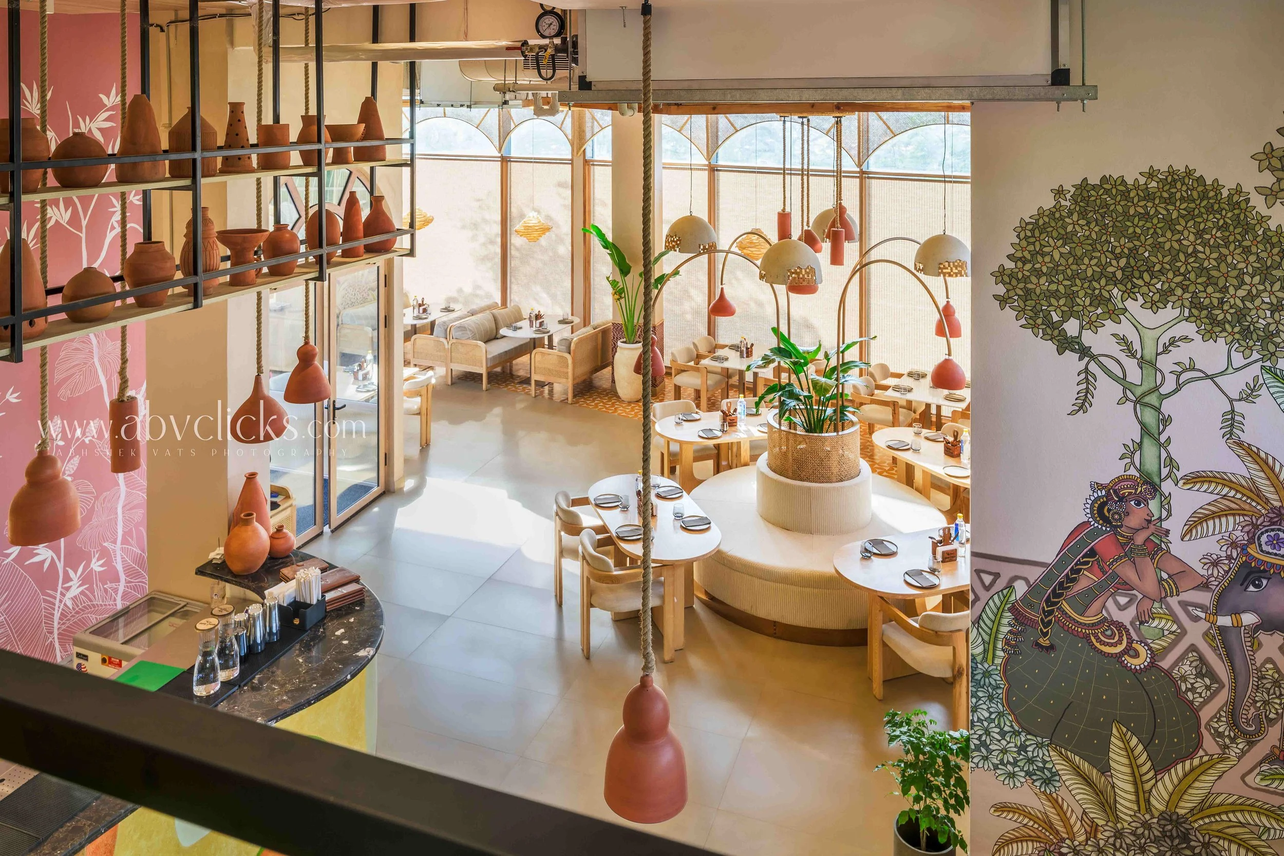 Interior view of a restaurant with tables set for dining, large windows letting in natural light, hanging pendant lights, green plants, and decorative pottery displayed on shelves, with colorful wall murals featuring a tree and traditional art.