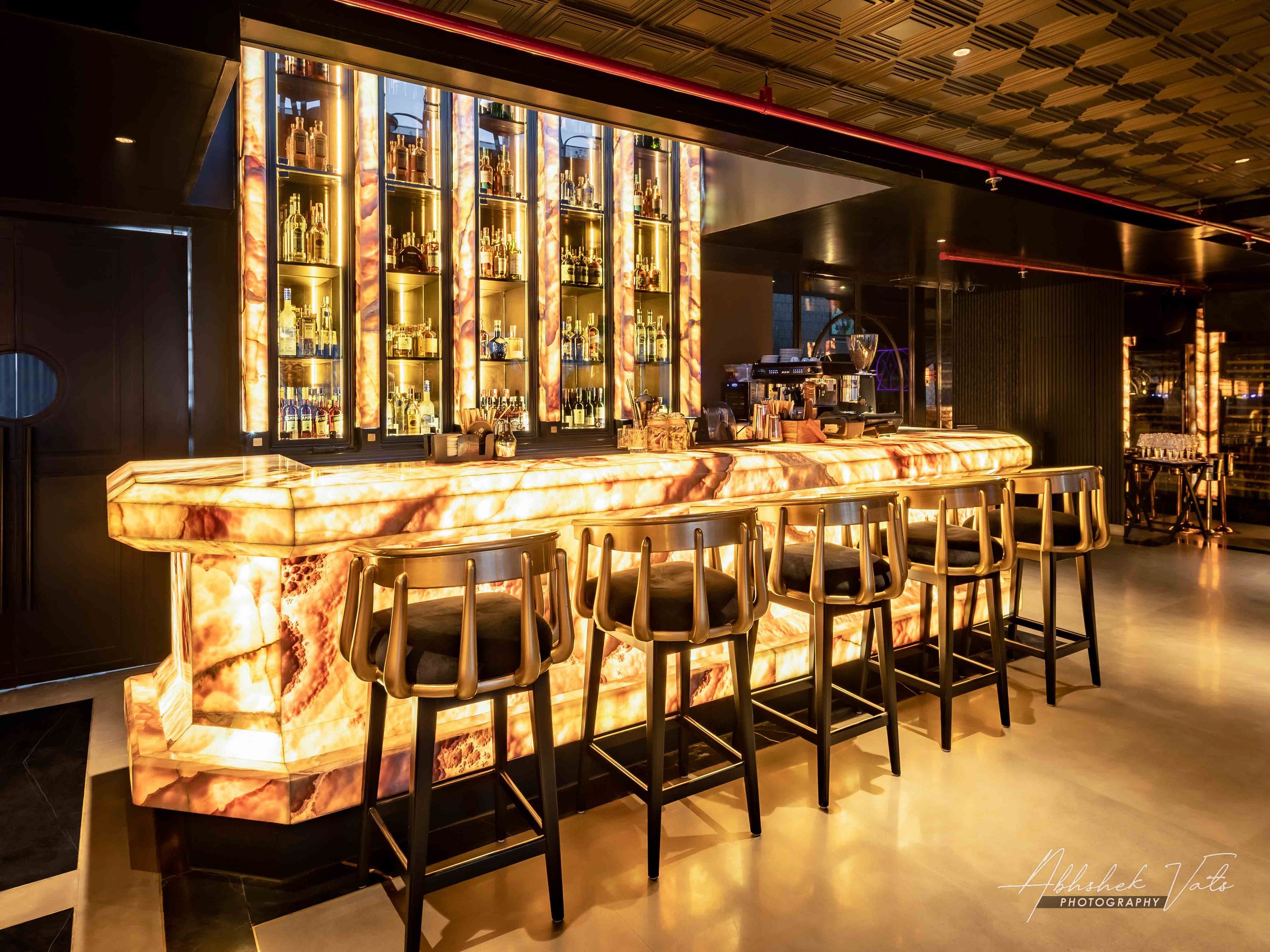 Bar counter with illuminated onyx surface, bottles of alcohol displayed on glass shelves behind, bar stools in front, modern design with warm lighting.