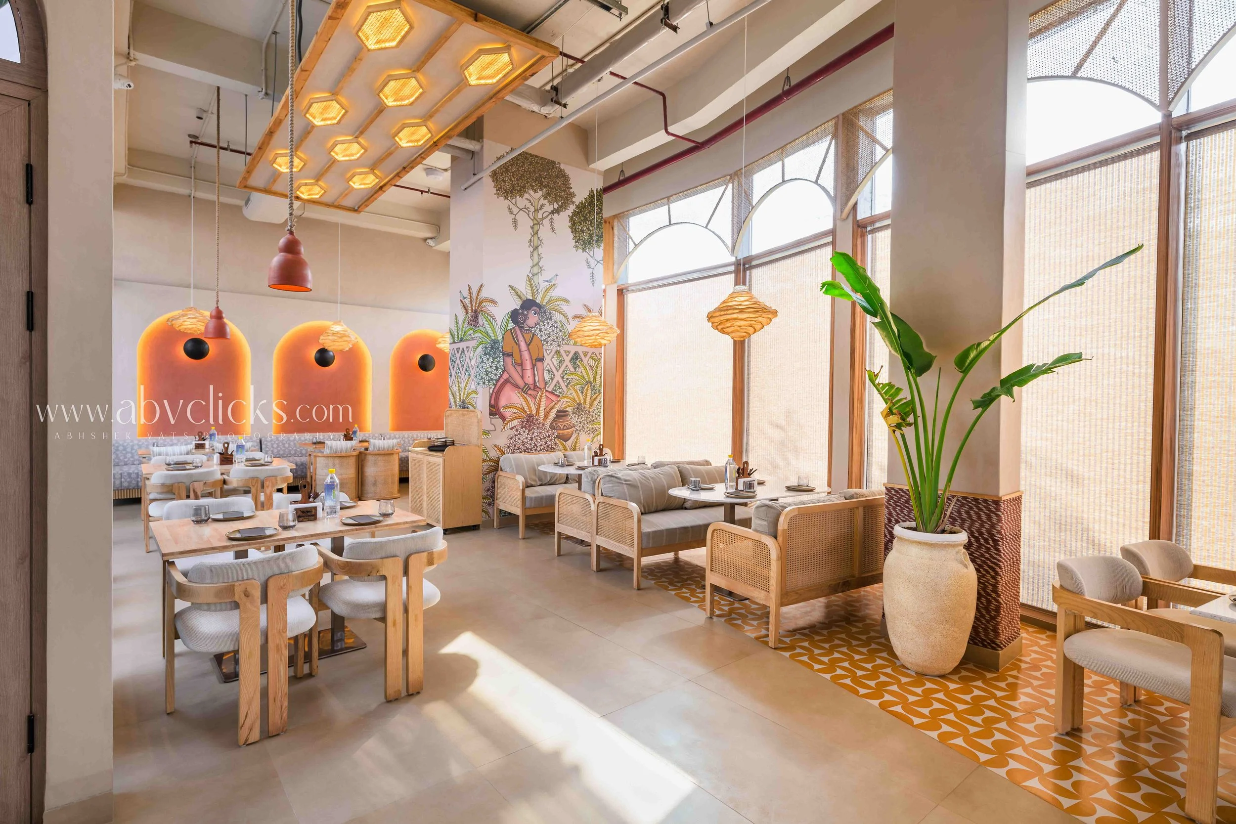 Interior of a bright restaurant with wooden furniture, large windows with blinds, decorative wall art, hanging light fixtures, and a large potted plant.