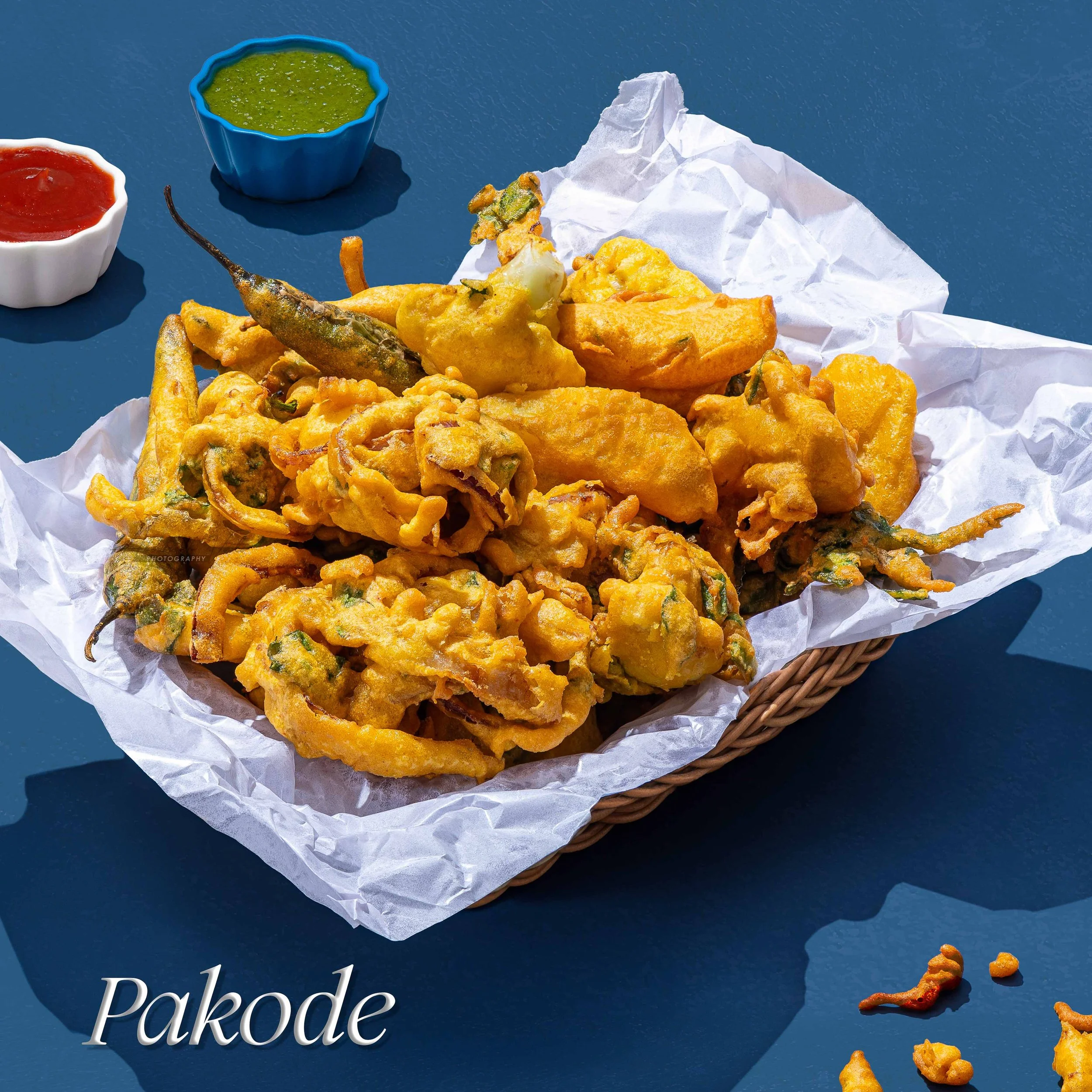 A basket with fried pakora and two small bowls of dipping sauces on a blue surface. One bowl contains red sauce and the other green sauce.