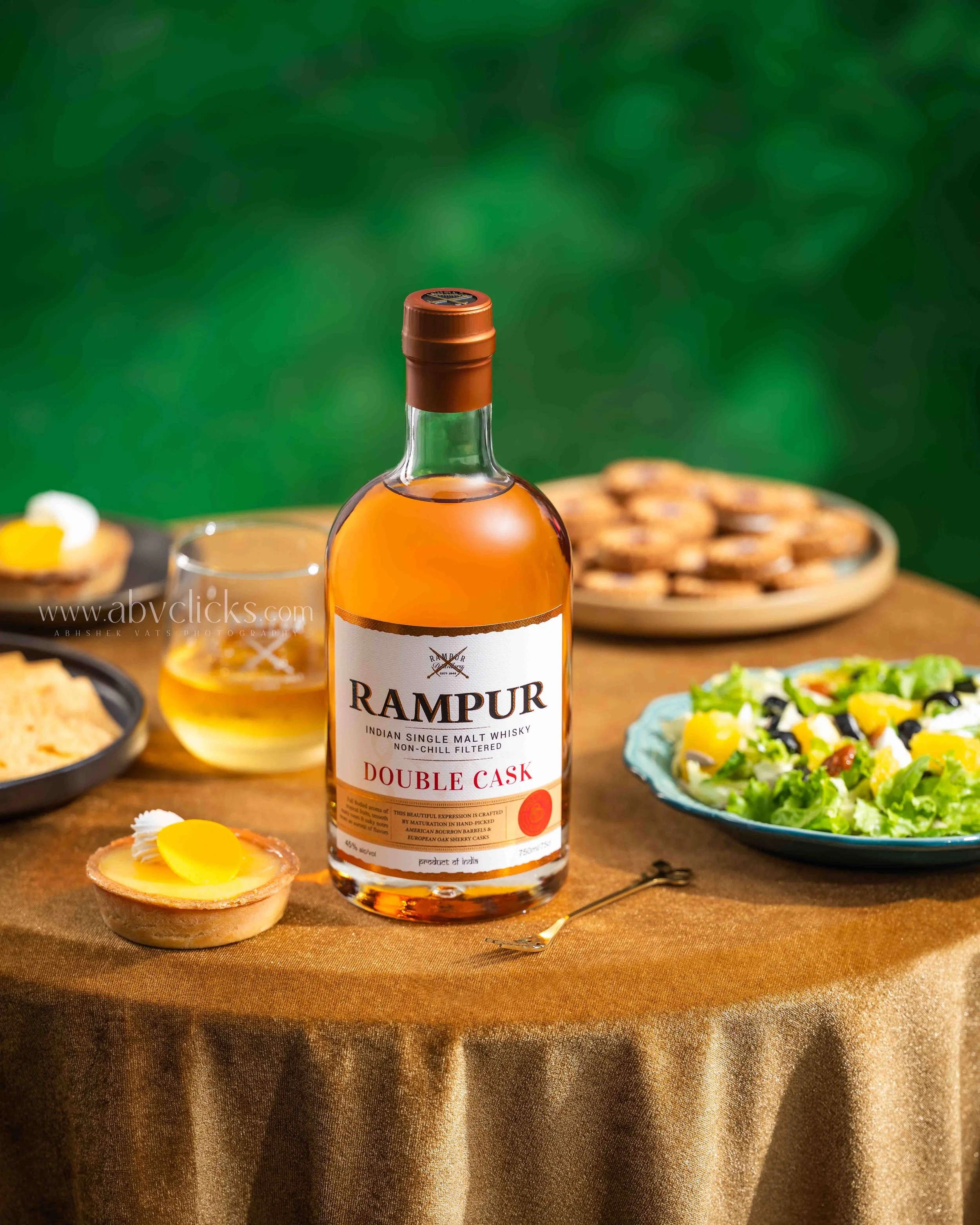 A bottle of Rampur Double Cask Indian single malt whisky on a table with food items in the background, including a salad and small tarts, on a brown tablecloth with a green blurred background.