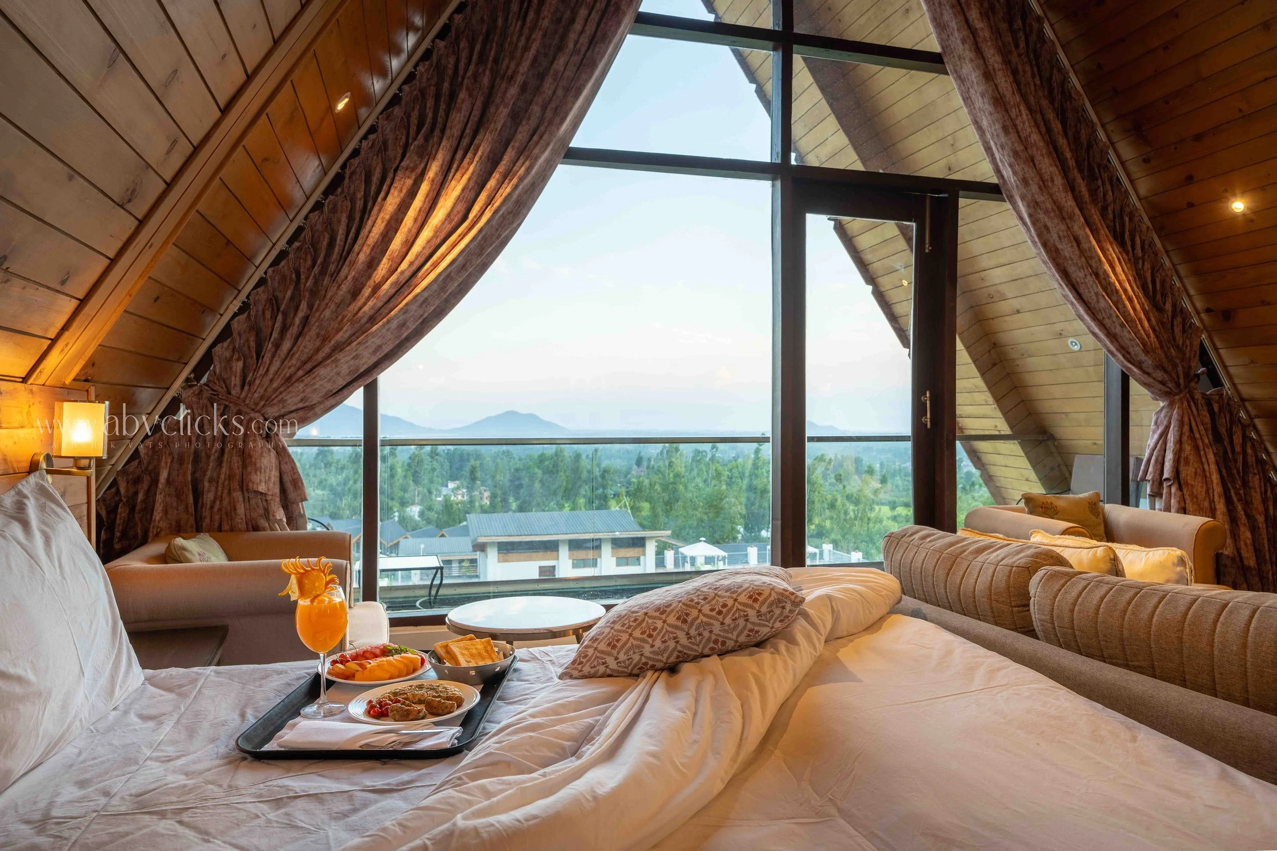 A cozy bedroom with large floor-to-ceiling windows showing a view of greenery and mountains. The room has wooden walls and ceiling, with curtains drawn partly aside. A bed with white sheets and a breakfast tray with food and drinks is in the foregrou
