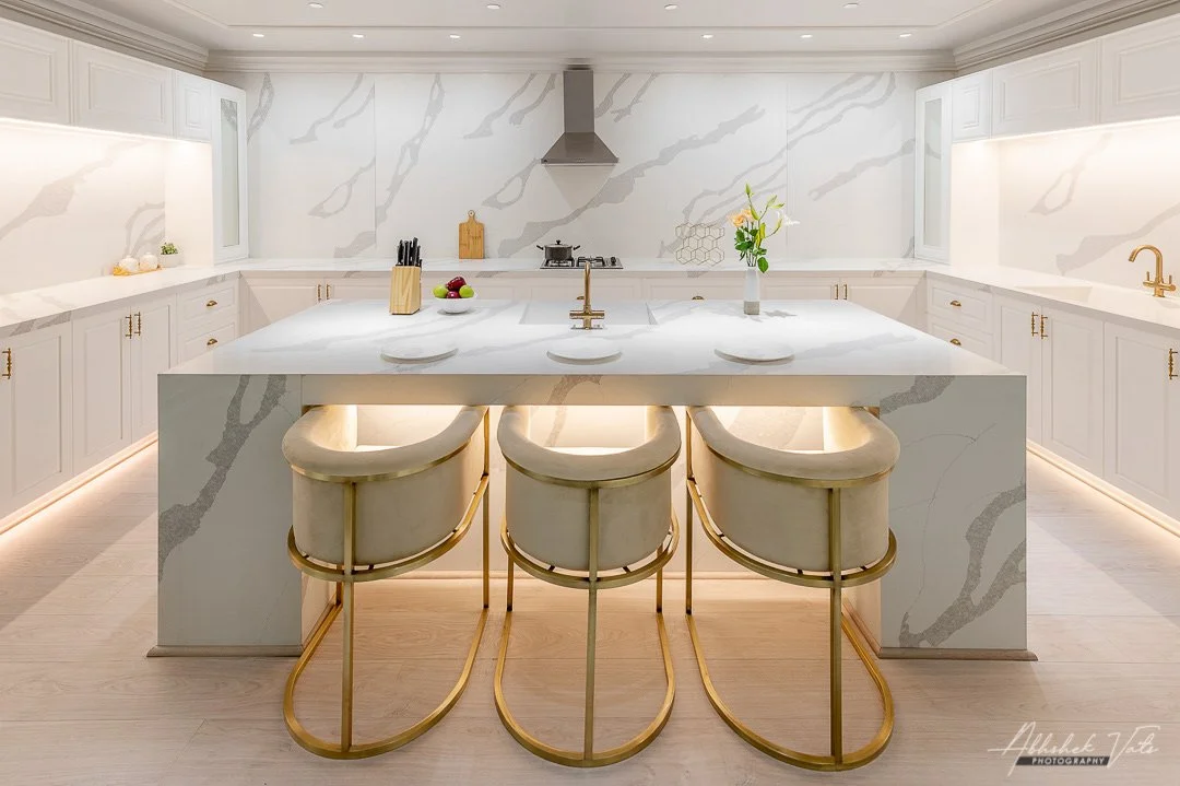 Modern white kitchen with marble countertops and a large island, decorated with a vase of flowers, bowls of fruit, and three bar stools with gold frames.
