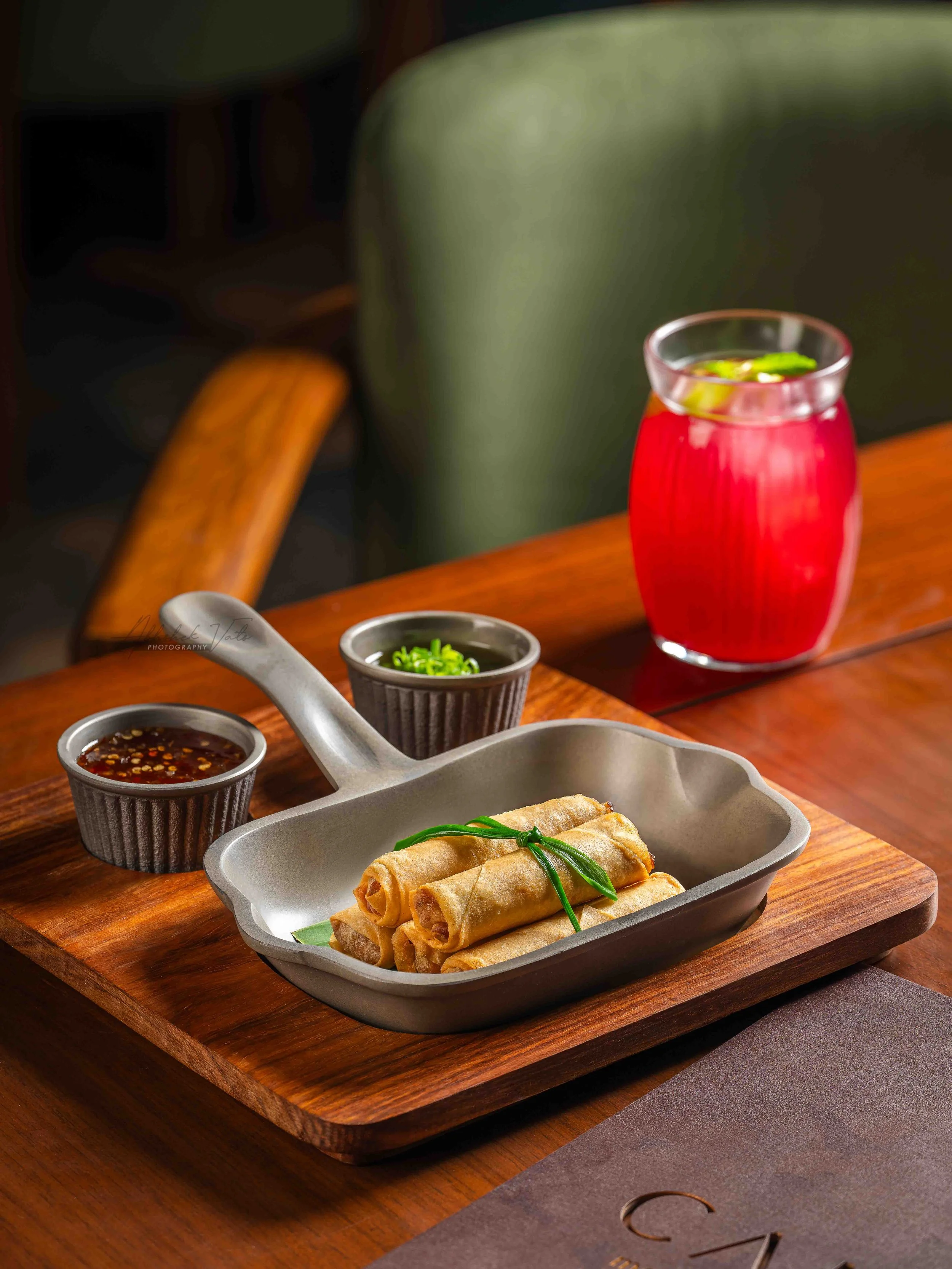 A wooden tray with spring rolls garnished with green herbs, accompanied by two small bowls of dipping sauces, and a glass of red drink with ice and lime slices.