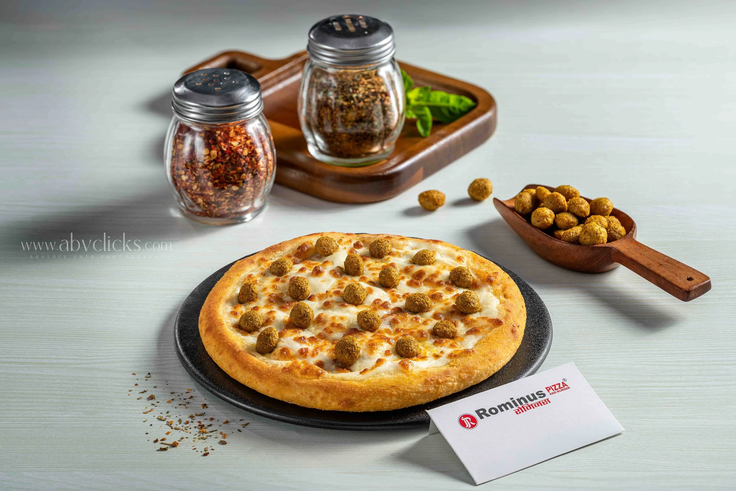 A pizza with cheese and small round sausage toppings on a black plate, surrounded by a container of spices, a small bowl of fried balls, and a menu card on a light-colored table.