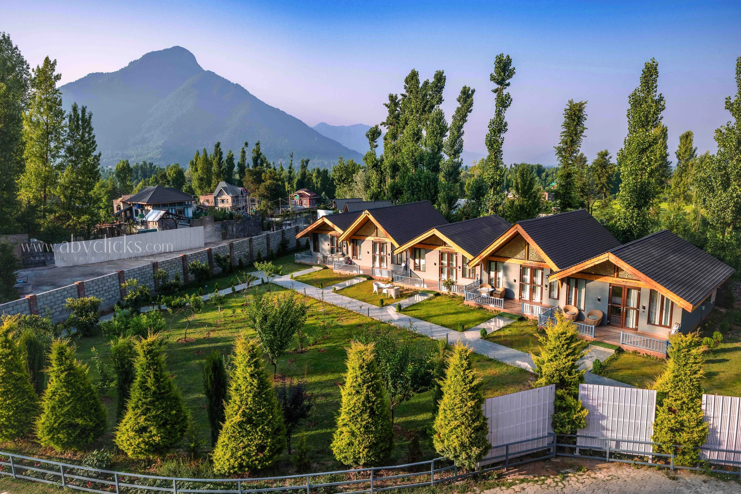 A scenic view of a small residential area with modern houses, surrounded by lush green trees and mountain hills in the background. Clear blue sky.