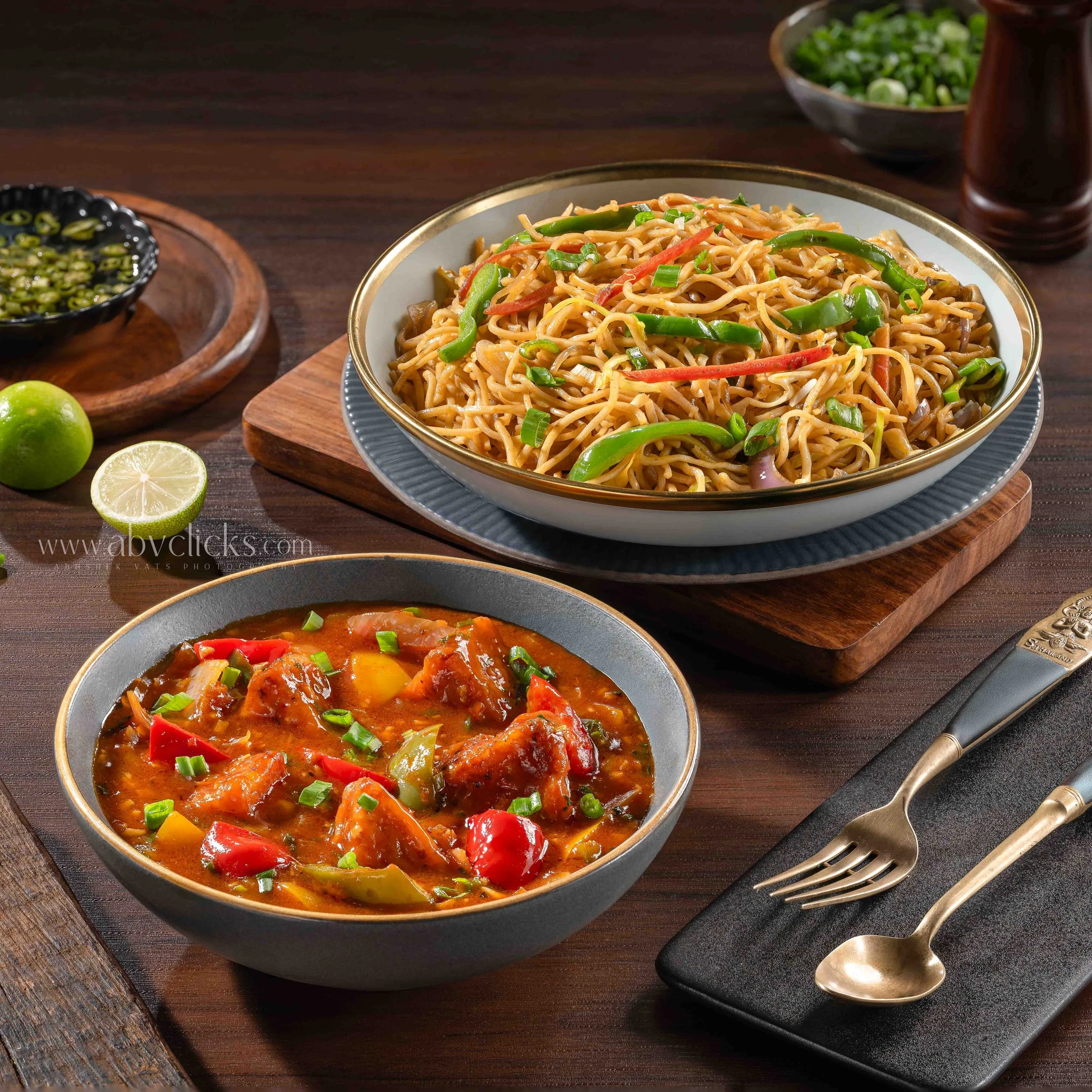 A bowl of stir-fried noodles with green and red bell peppers and chopped green onions on a wooden table, with a bowl of chopped green onions in the background and a lime and lemon halves in the foreground.