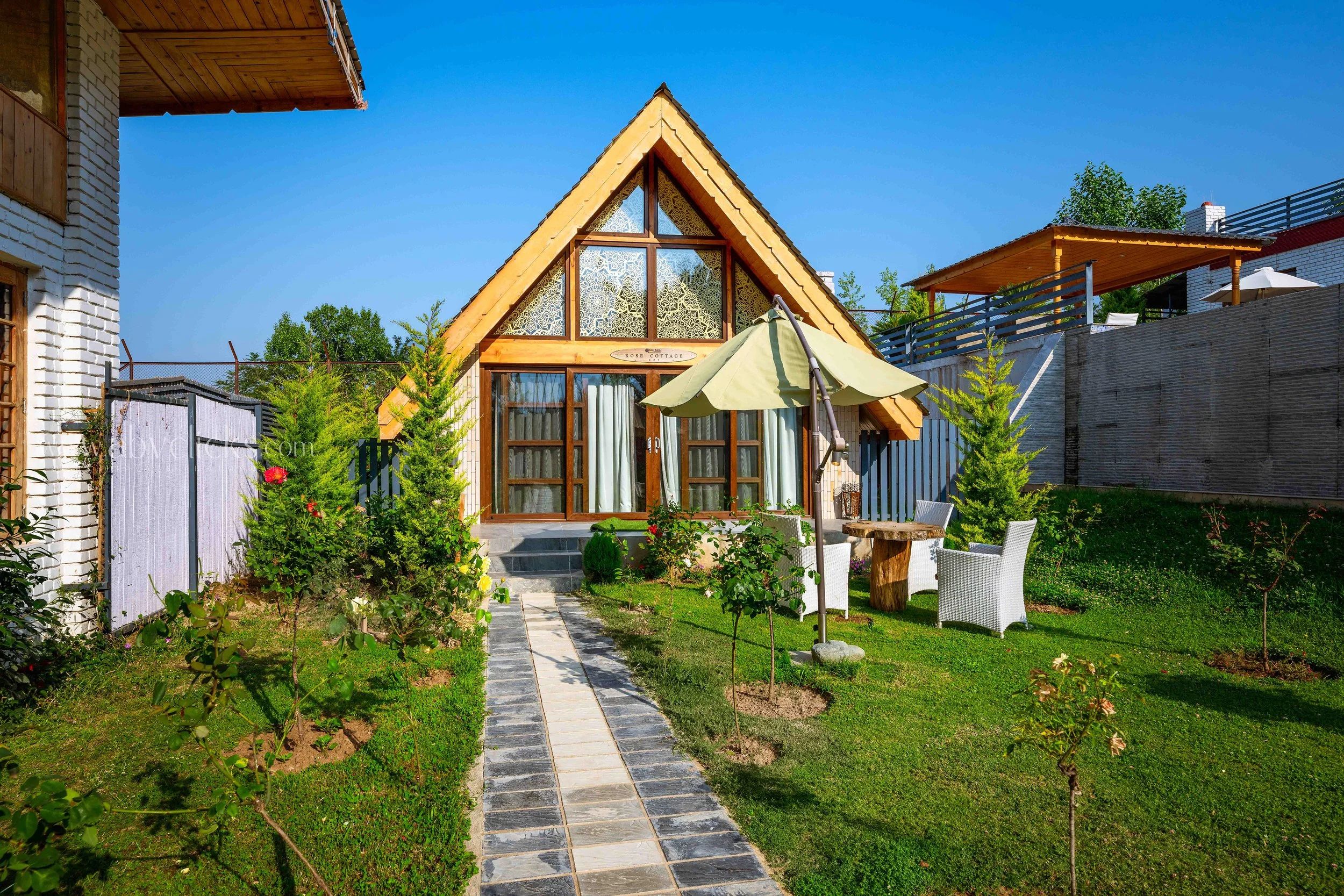 A charming A-frame cottage with glass windows and a wooden door, situated in a lush garden with a stone pathway, umbrellas, and outdoor seating, under a clear blue sky.