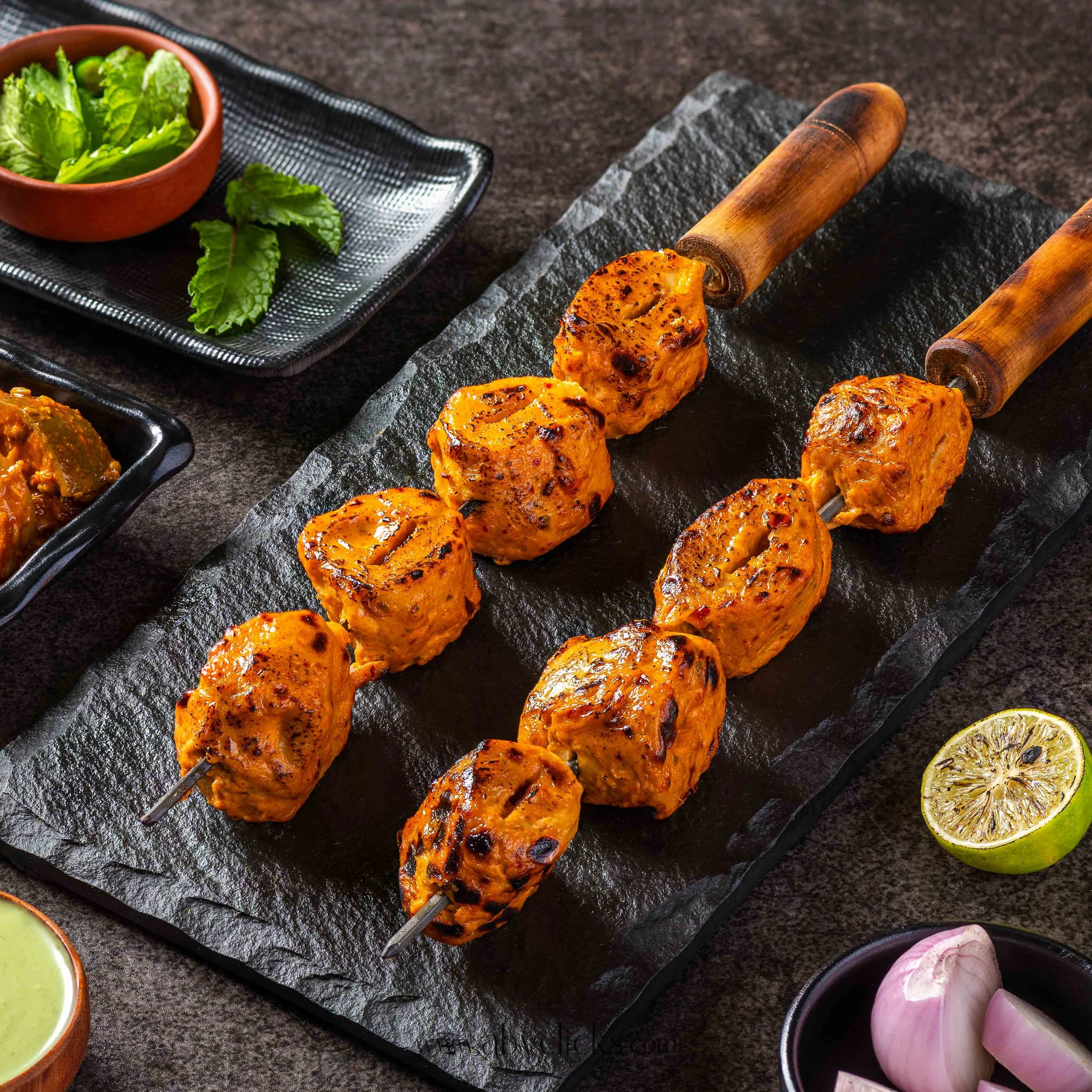 Skewers of grilled chicken tikka served on a black slate platter, with lime slices and a side of fresh green lettuce in a terracotta bowl, garnish with lemon slices and red onions